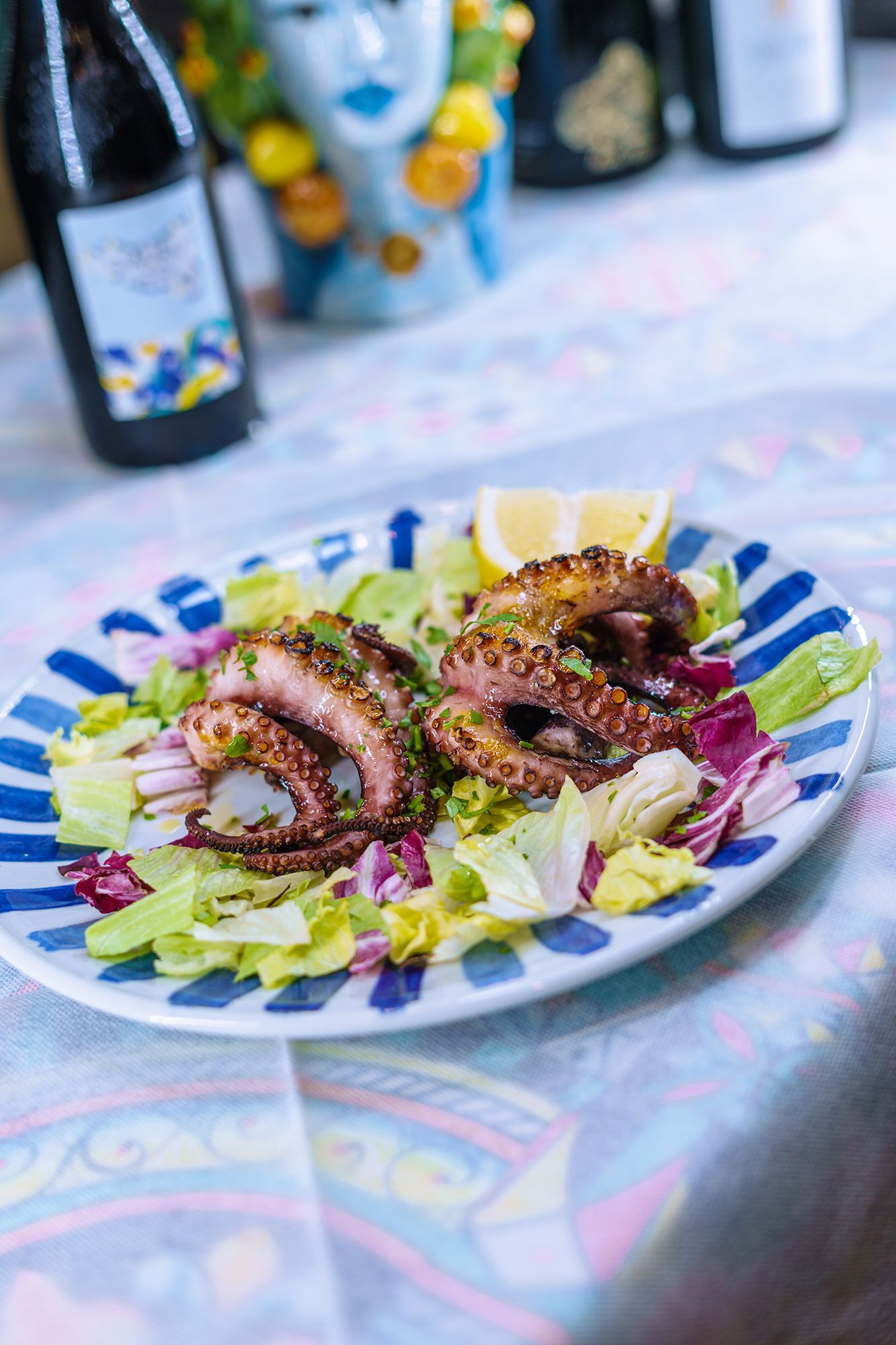 Polpo grigliato su un piatto a righe con misticanza e uno spicchio di limone, su un tavolo con bottiglie di vino.