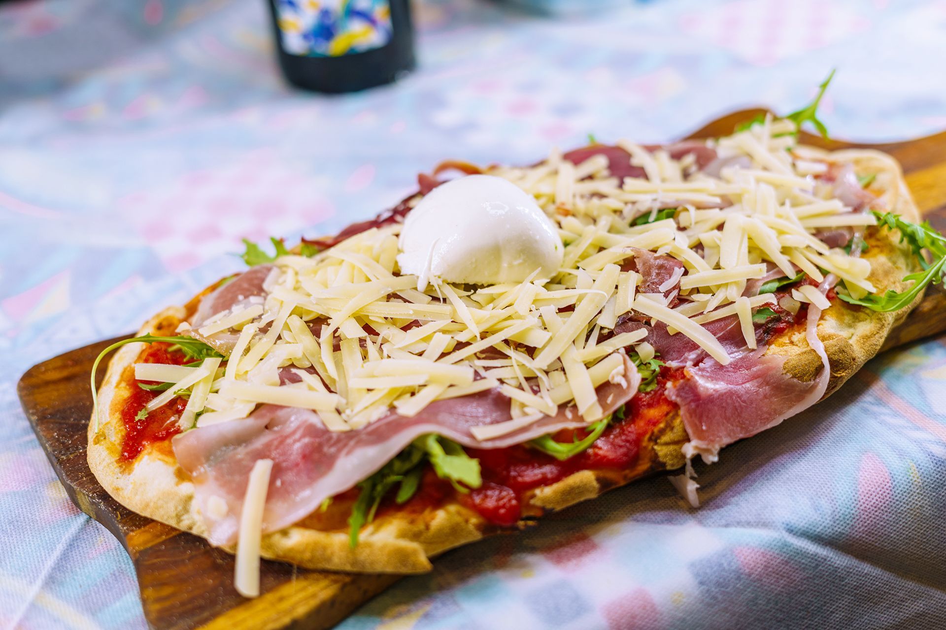 Pizza piatta con salsa di pomodoro, prosciutto, rucola, formaggio grattugiato e una pallina di mozzarella su un tagliere di legno.