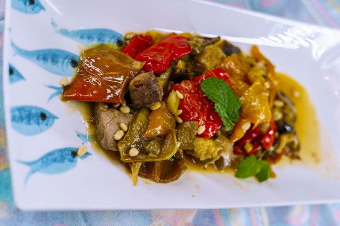Misto di verdure con peperoni rossi, arancioni e verdi, su un piatto bianco decorato con pesci azzurri, guarnito con menta e pinoli.