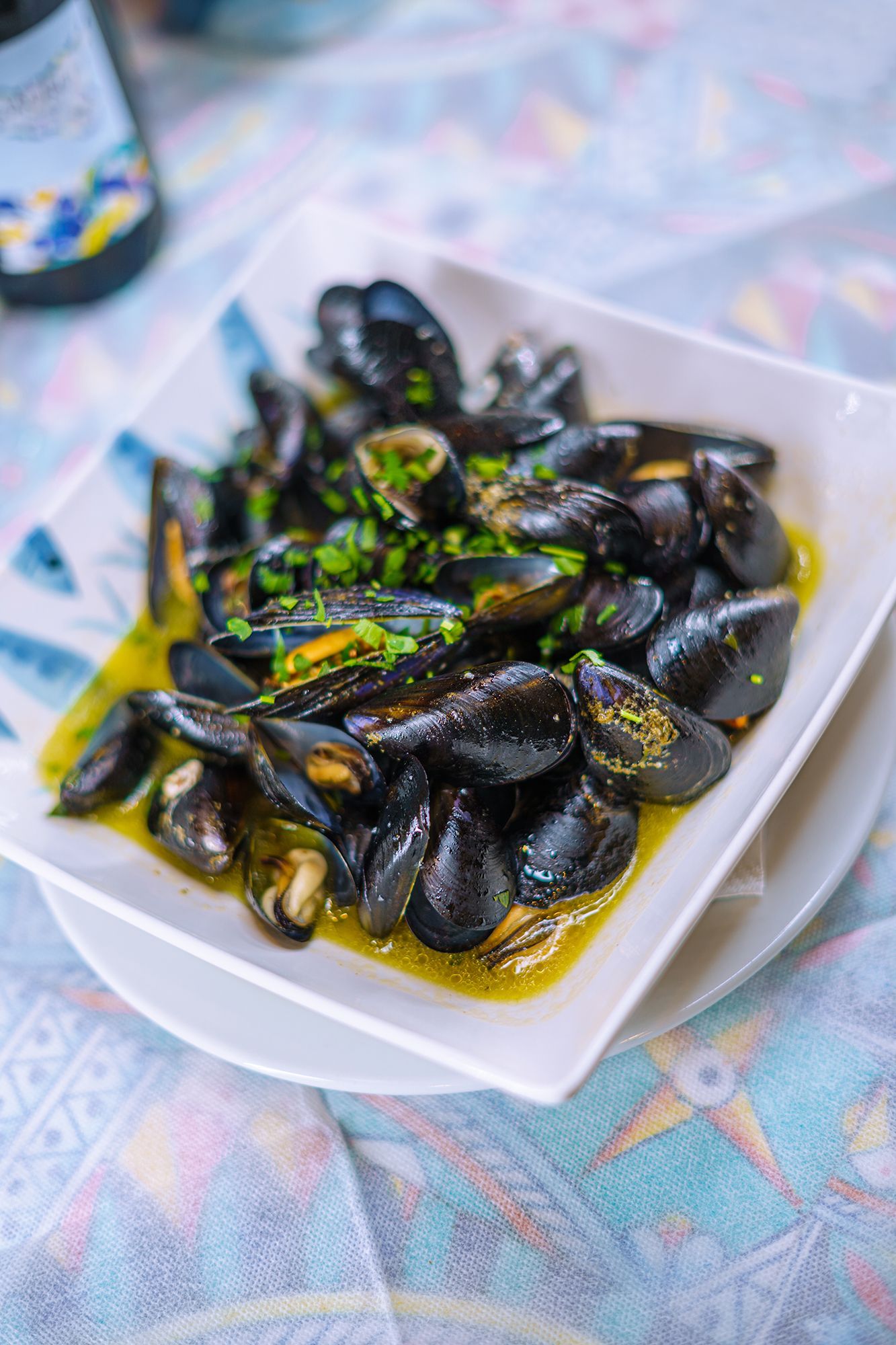Cozze al vapore in brodo, guarnite con erbe verdi, servite in una ciotola quadrata bianca su un tavolo.