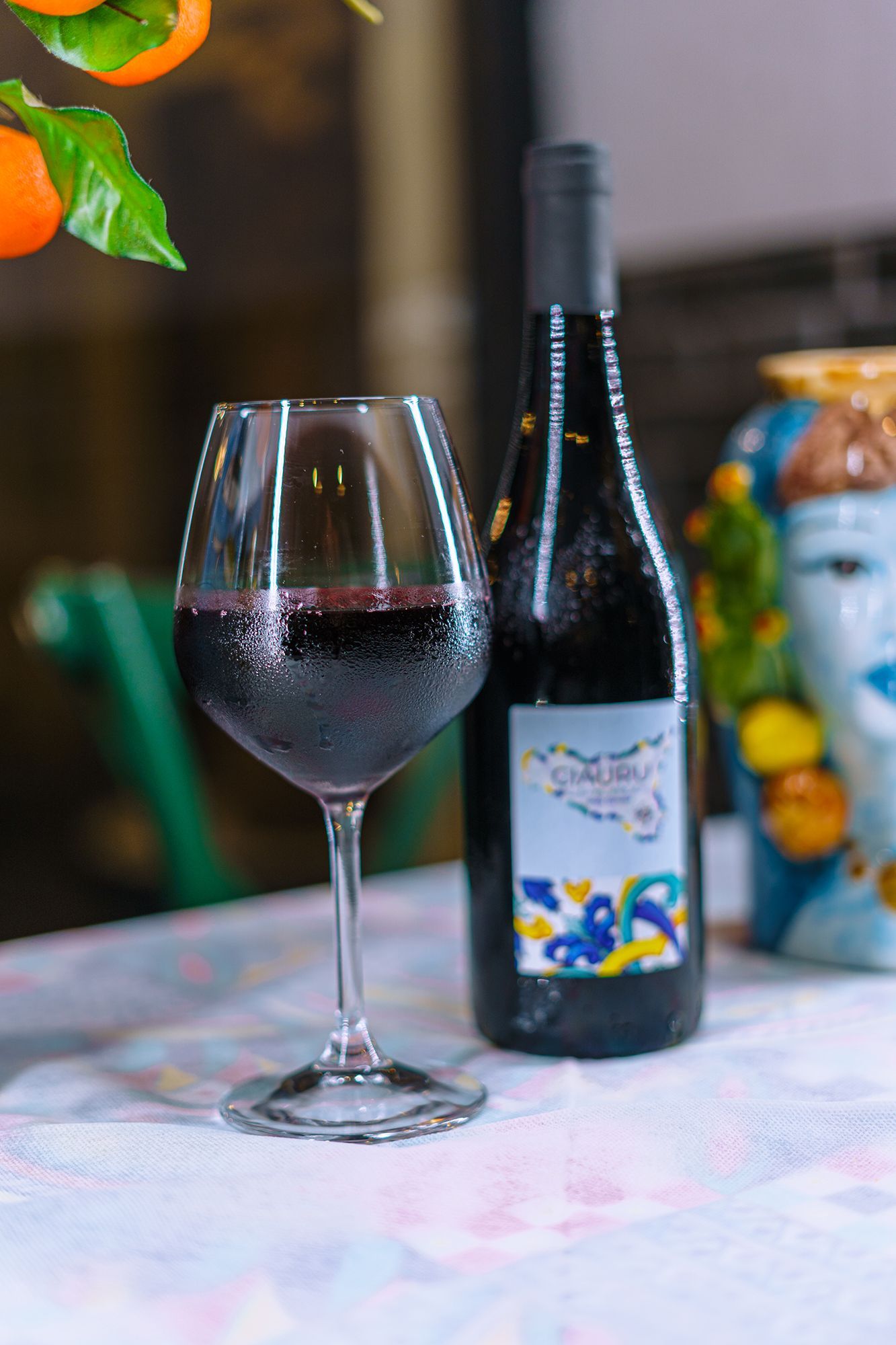 Un bicchiere di vino rosso e una bottiglia sono appoggiati su un tavolo, con arance e una testa di ceramica sullo sfondo.