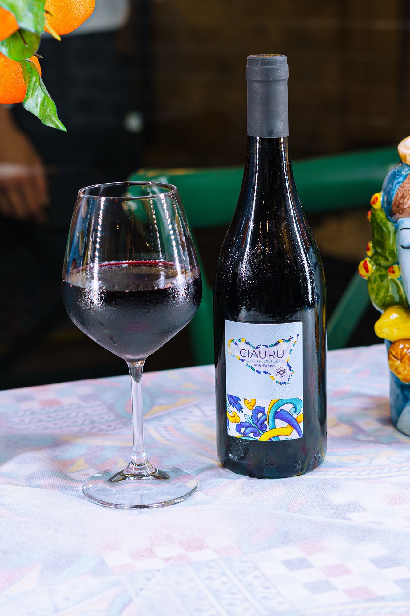Bottiglia di vino rosso e bicchiere pieno su un tavolo con un'etichetta colorata, arance e figure decorative.