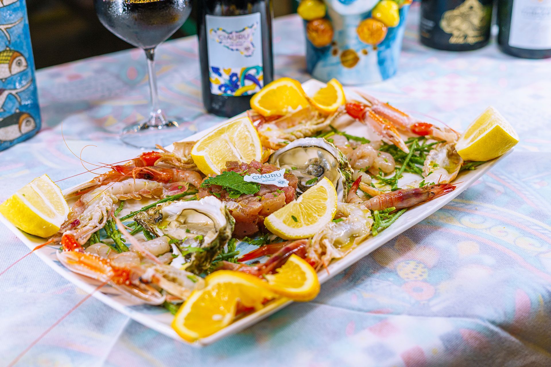 Piatto di frutti di mare con limoni e arance su un tavolo; bottiglie e bicchiere di vino sullo sfondo.
