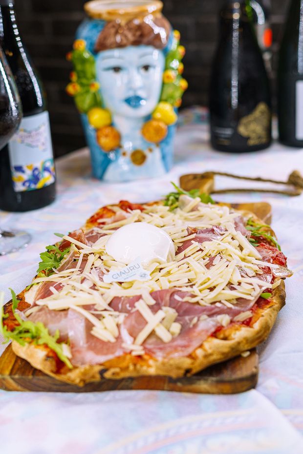 Pizza con prosciutto, formaggio, rucola e una cucchiaiata di formaggio bianco, su un tagliere di legno, con una testa decorativa sullo sfondo.