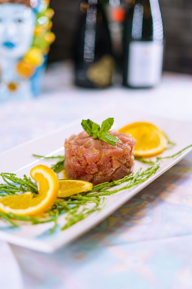 Tartare di tonno su piatto bianco, guarnita con fette d'arancia, verde e menta, con bottiglie sullo sfondo.