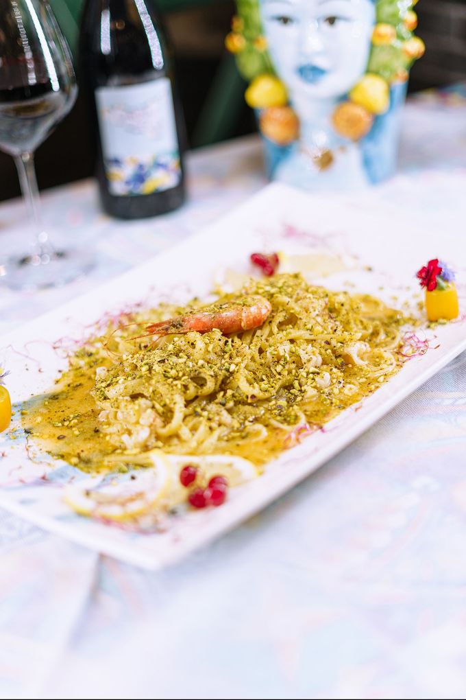 Piatto di pasta con gamberi e pistacchi su un piatto rettangolare bianco, con un bicchiere di vino e una bottiglia sullo sfondo.