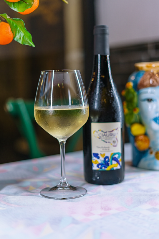 Un bicchiere di vino bianco accanto a una bottiglia, su un tavolo con motivi floreali, vicino a frutti arancioni.
