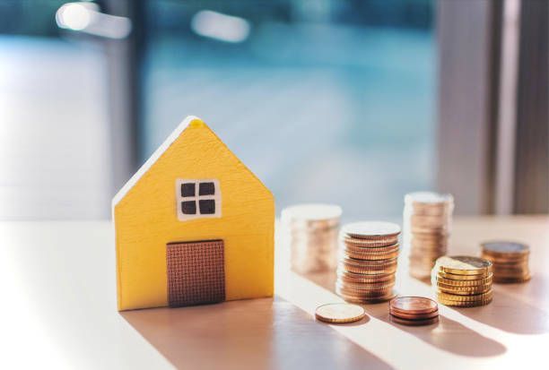 A small yellow house is sitting on a table next to stacks of coins.