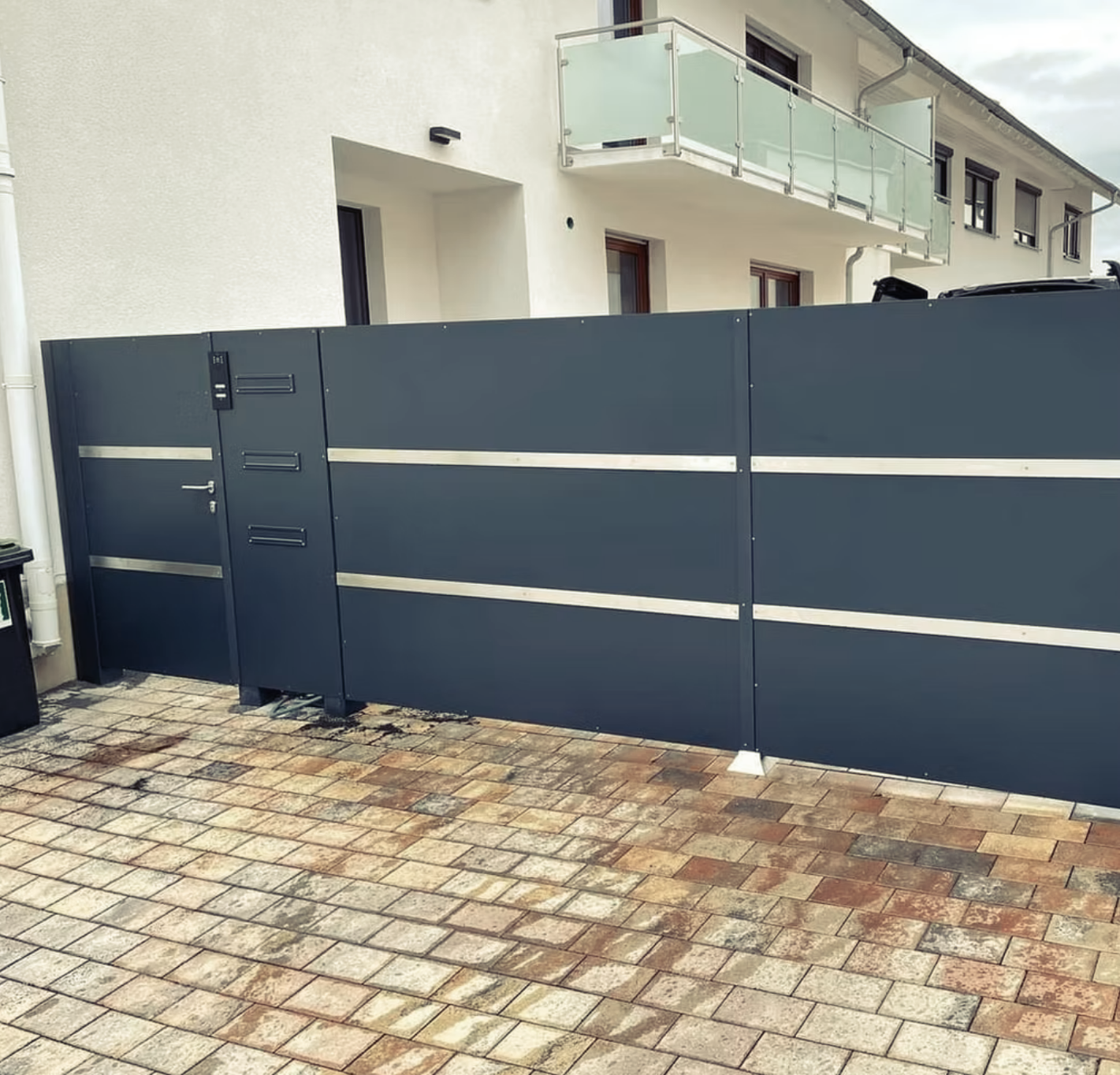 Ein schwarzer Zaun umgibt eine gepflasterte Auffahrt vor einem Haus.