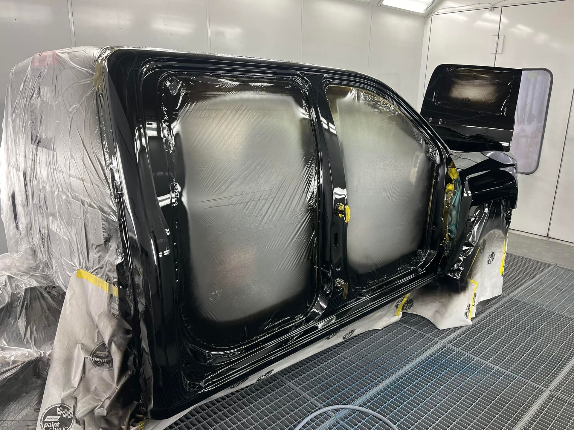 A black truck body prepped for painting in a spray booth, with doors and windows covered in plastic sheeting.