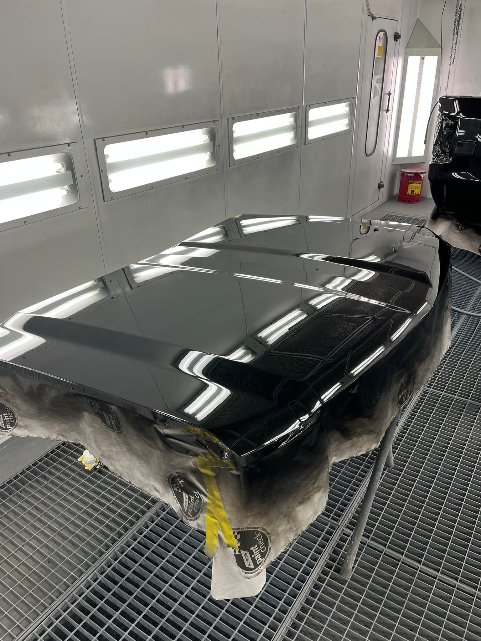 A freshly painted, glossy black car hood rests on a stand inside a brightly lit professional auto body spray booth.