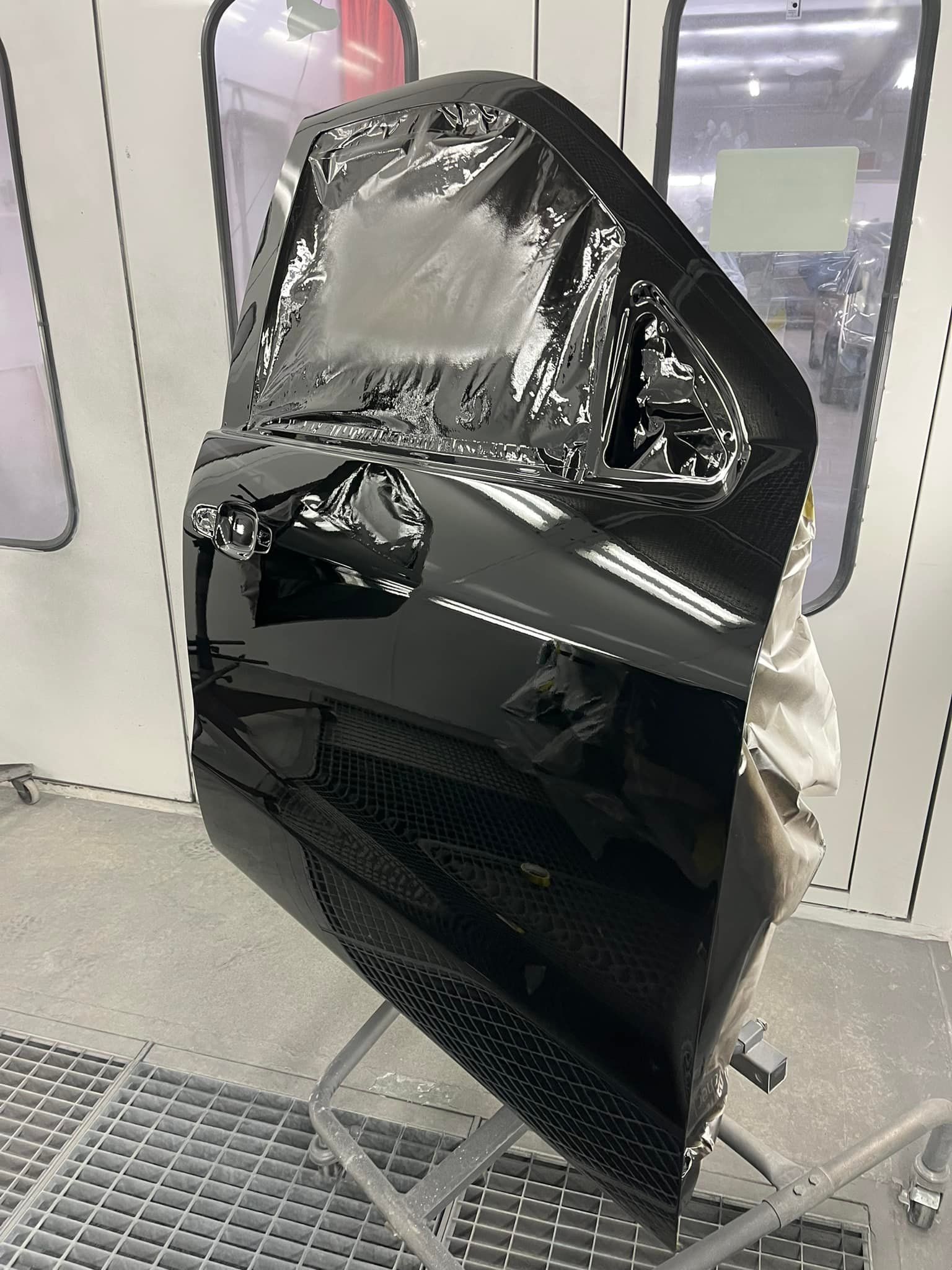 A black car door panel is mounted on a stand inside an auto body paint booth, with the window area covered in plastic.