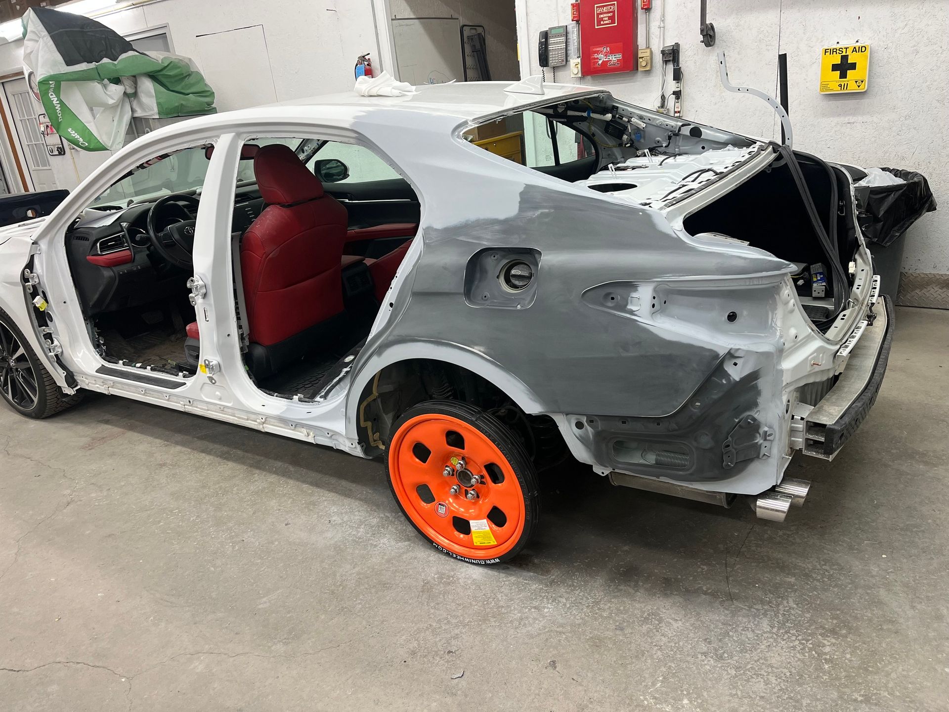 A white car in a garage with red seats and a bright orange spare tire, undergoing body repair on the rear panel.