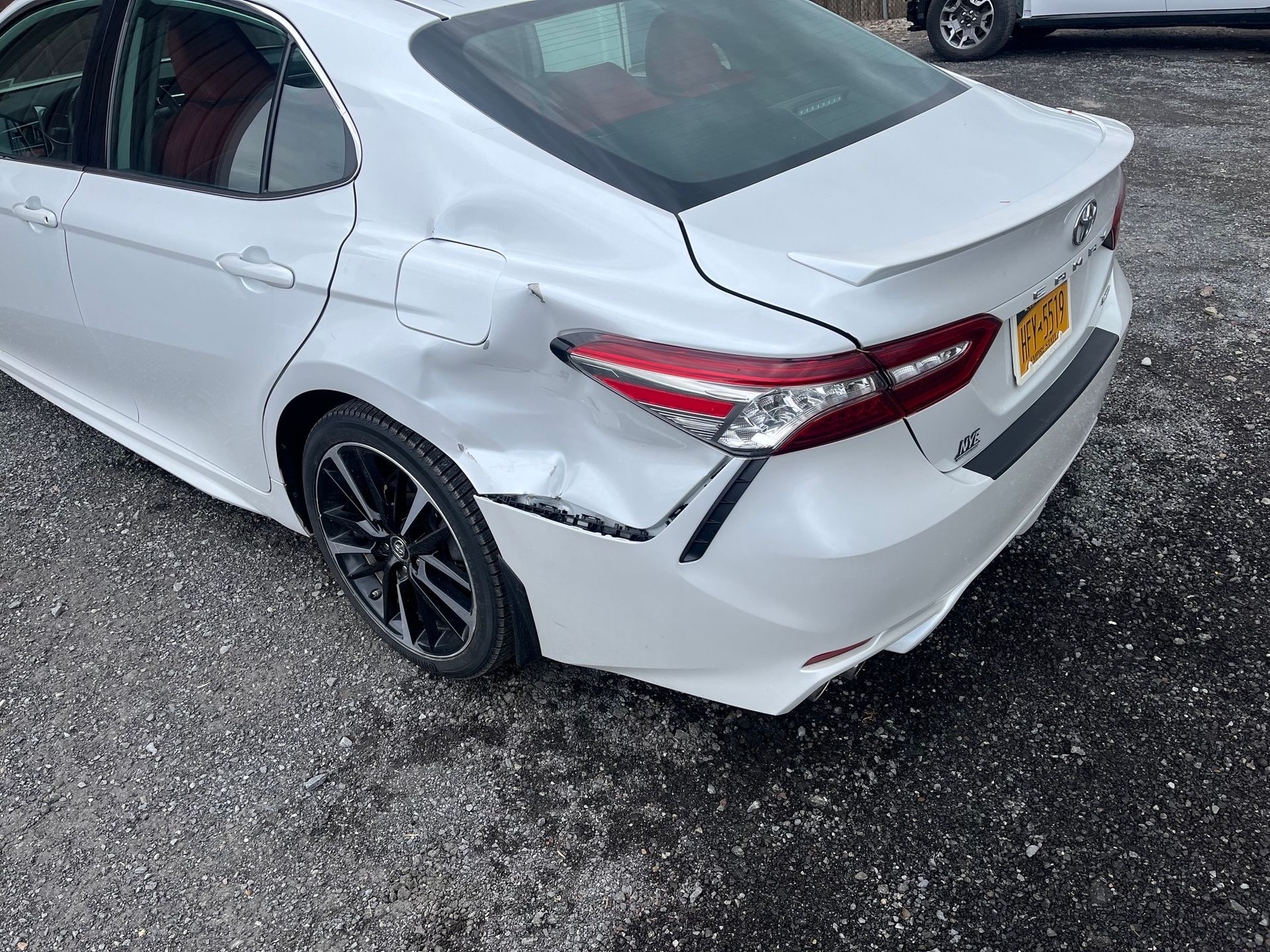White Toyota sedan with significant collision damage to the rear quarter panel and bumper, parked on a gravel lot.