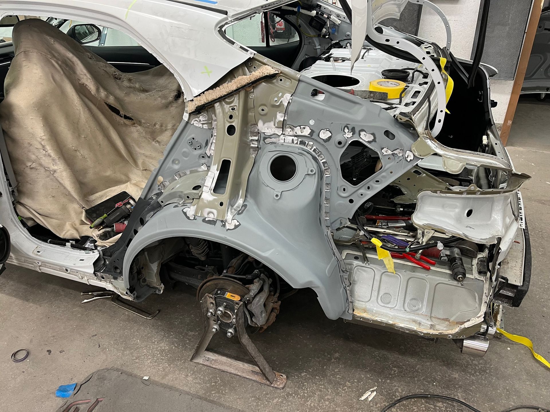 The rear section of a white car undergoing body repair in a shop, with the quarter panel and bumper removed.