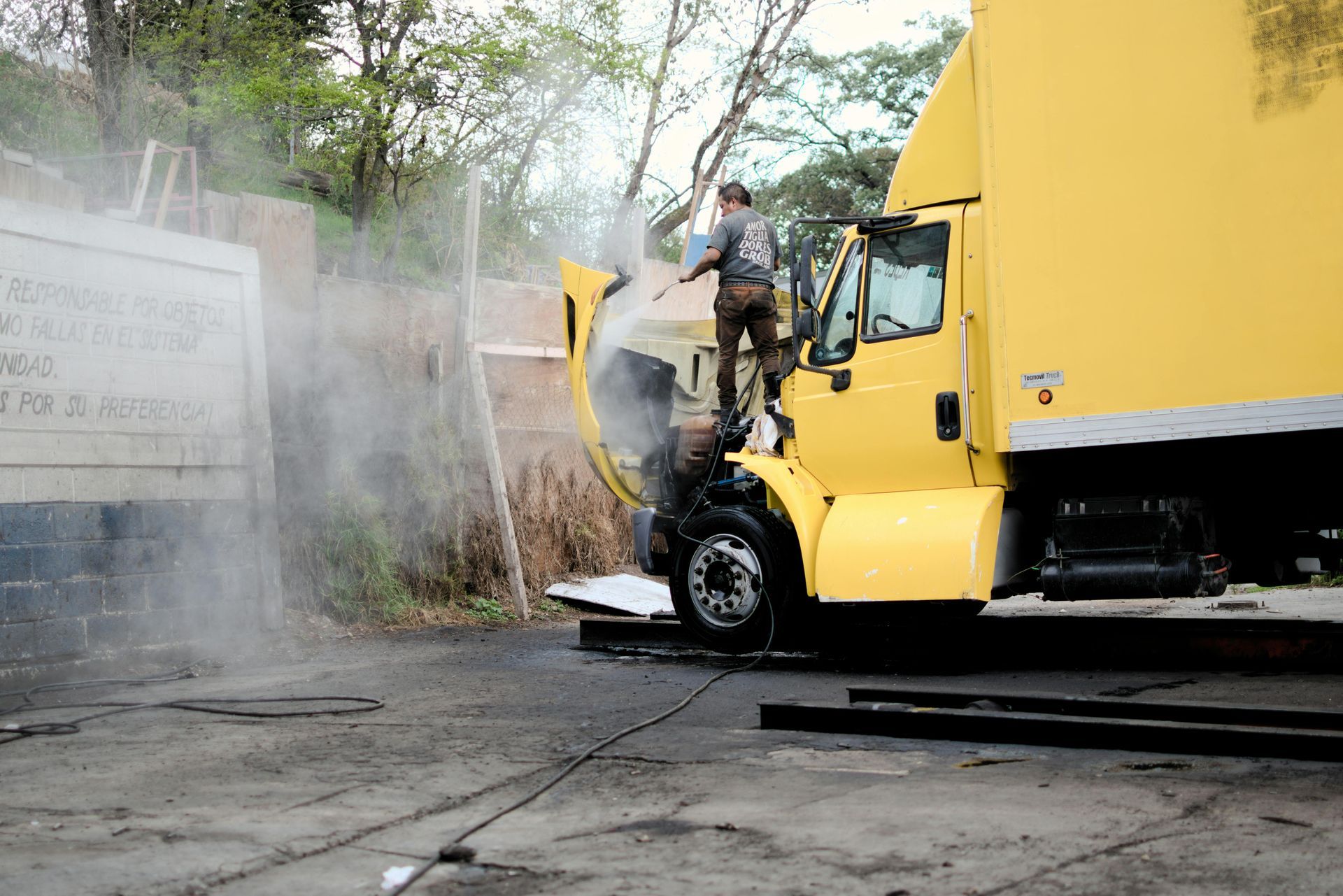 Man power-washes a yellow semi-truck engine. Gray water vapor surrounds them. Outdoors.