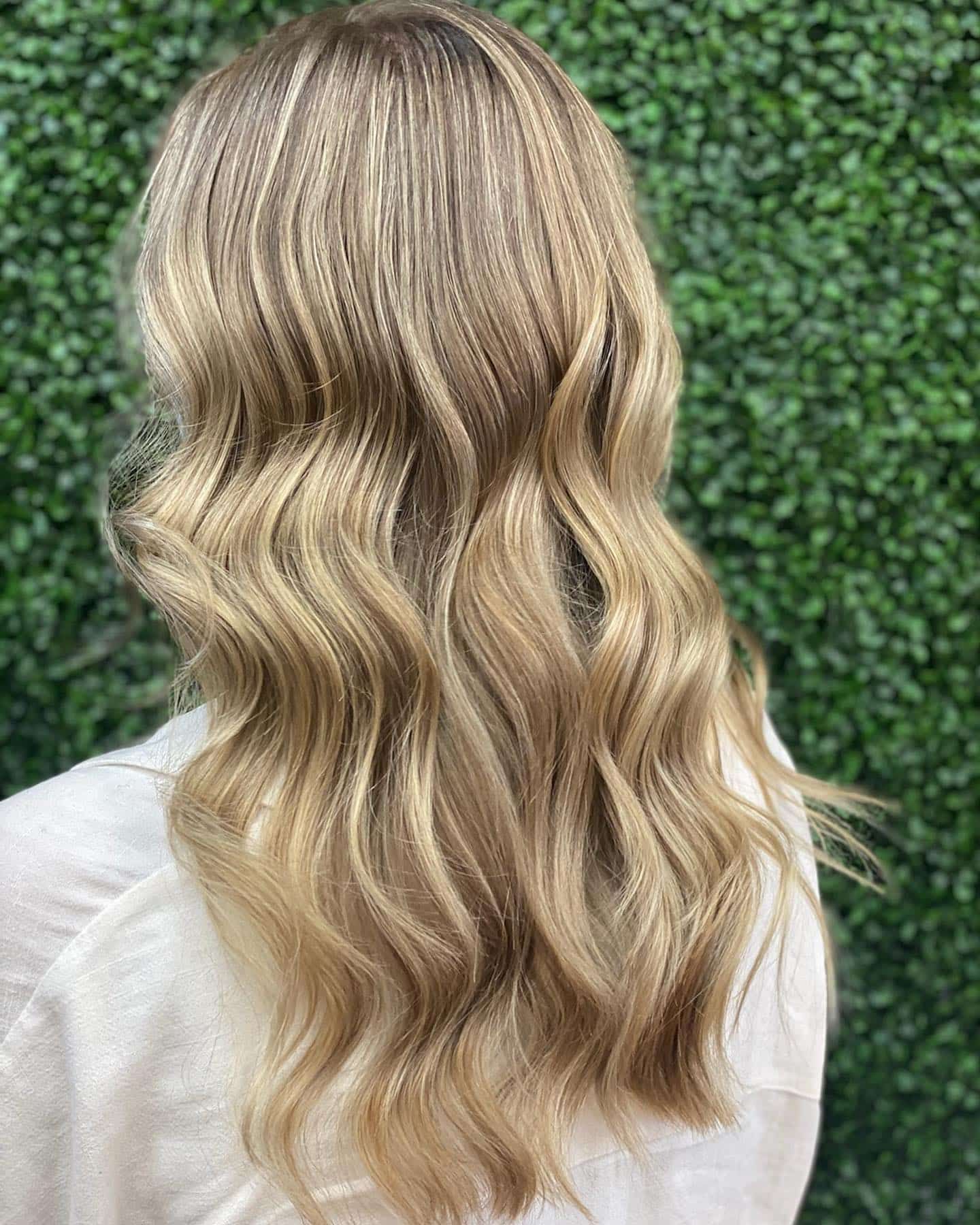 Woman With Long Wavy Blonde Hair, Standing in Front of a Green Hedge — Lavish Hair in Shepparton, VIC