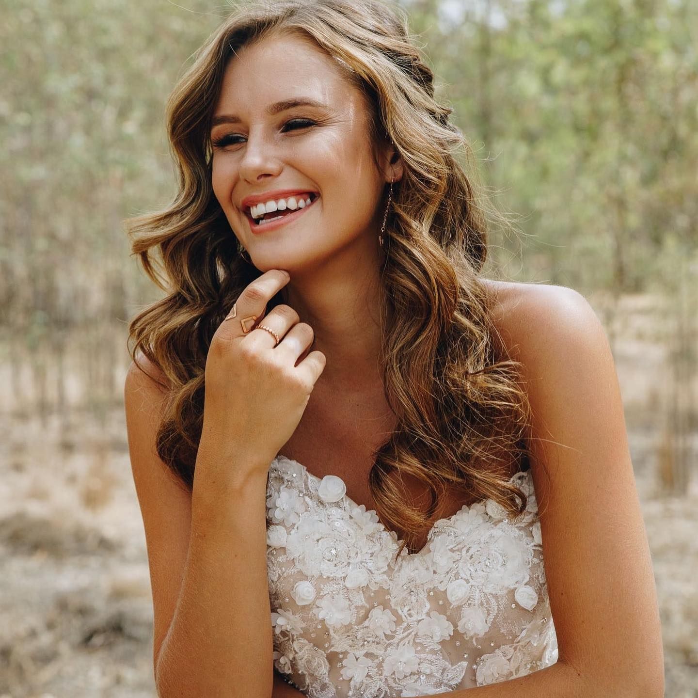 Woman With Wavy Brown Hair Smiles, Wearing a White, Floral-detailed Dress Outdoors — Lavish Hair in Shepparton, VIC