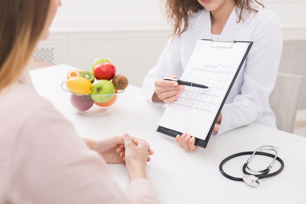 A Woman Is Sitting At A Table Talking To A Doctor Who Is Holding A Clipboard — Wegner Therapies In Pialba, QLD