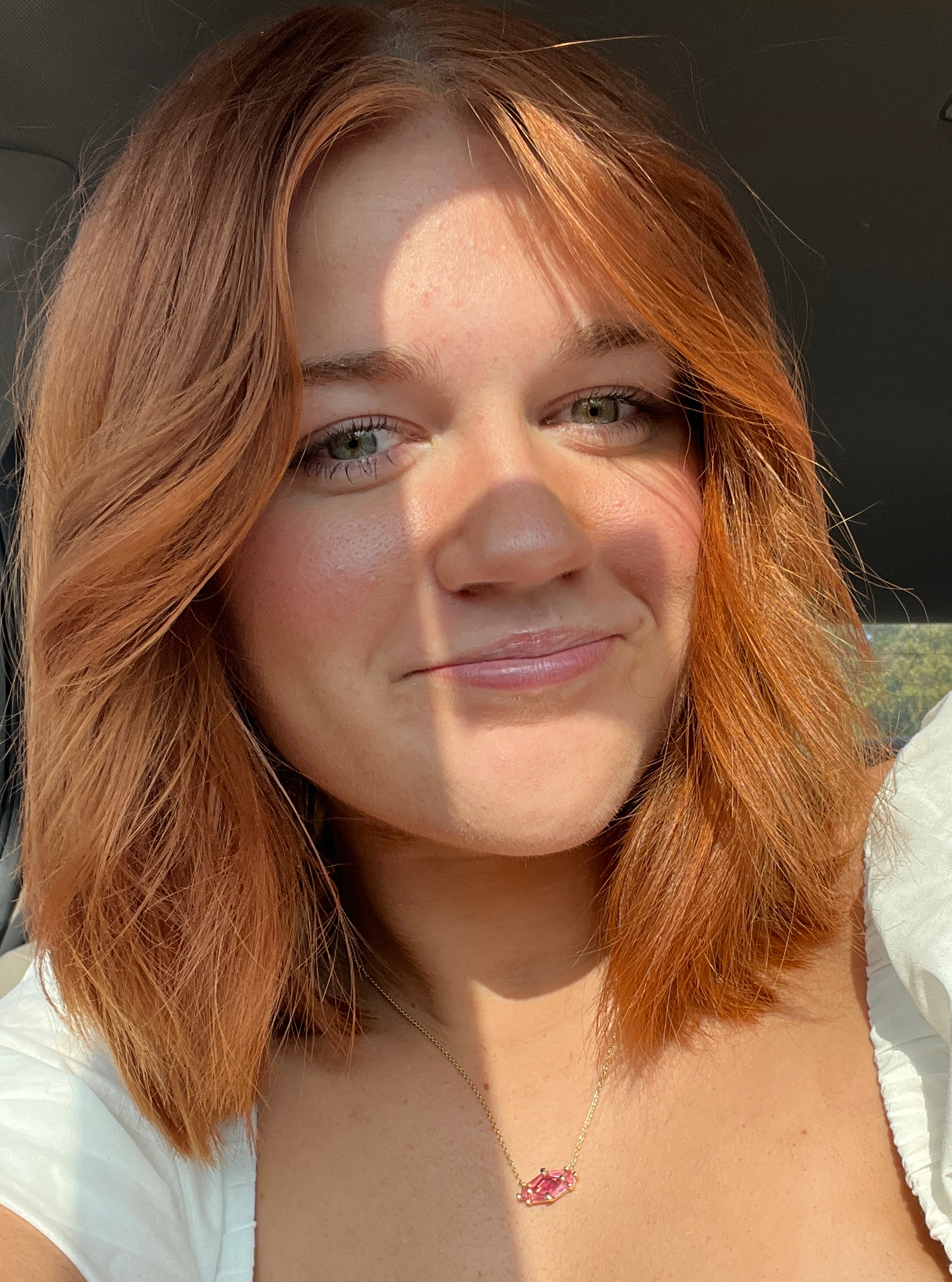 Woman with red hair smiling, wearing a white top and a necklace, lit by sunlight.