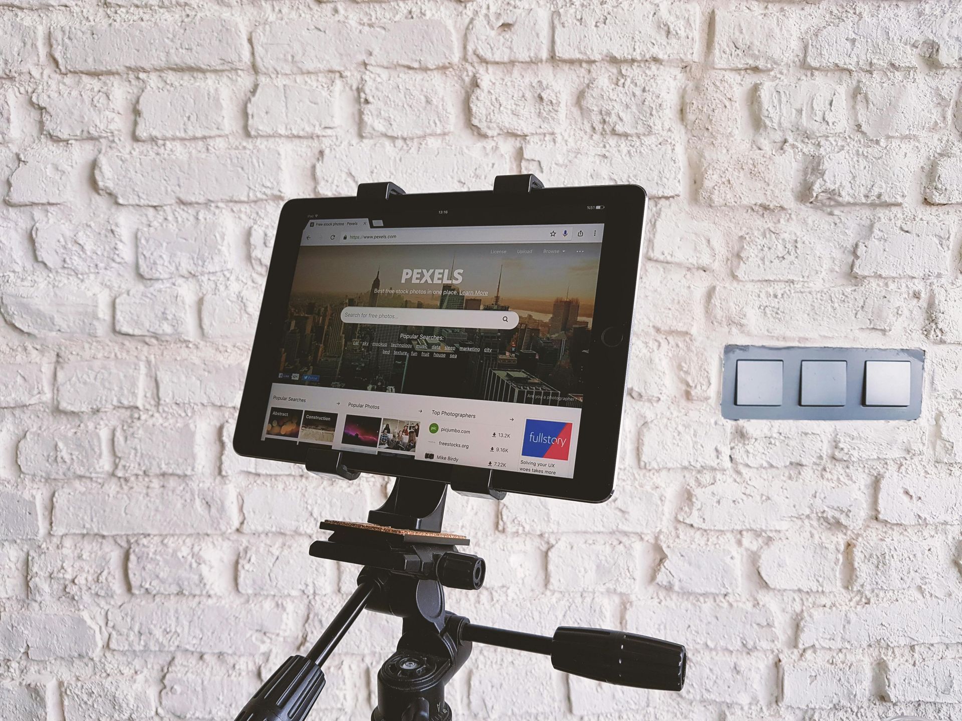 Tablet on tripod displaying a website, positioned in front of a white brick wall with a light switch.