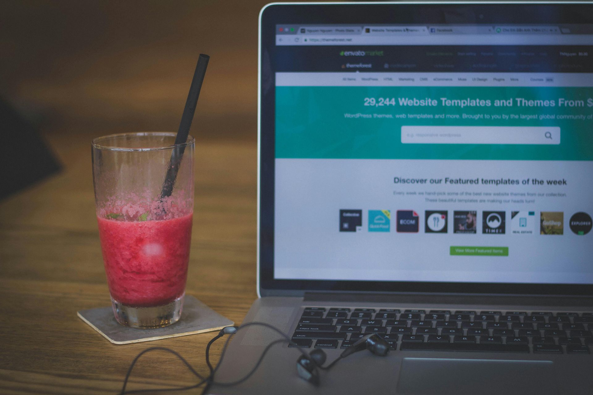 Laptop with website on screen, smoothie, and earbuds on a wooden surface.