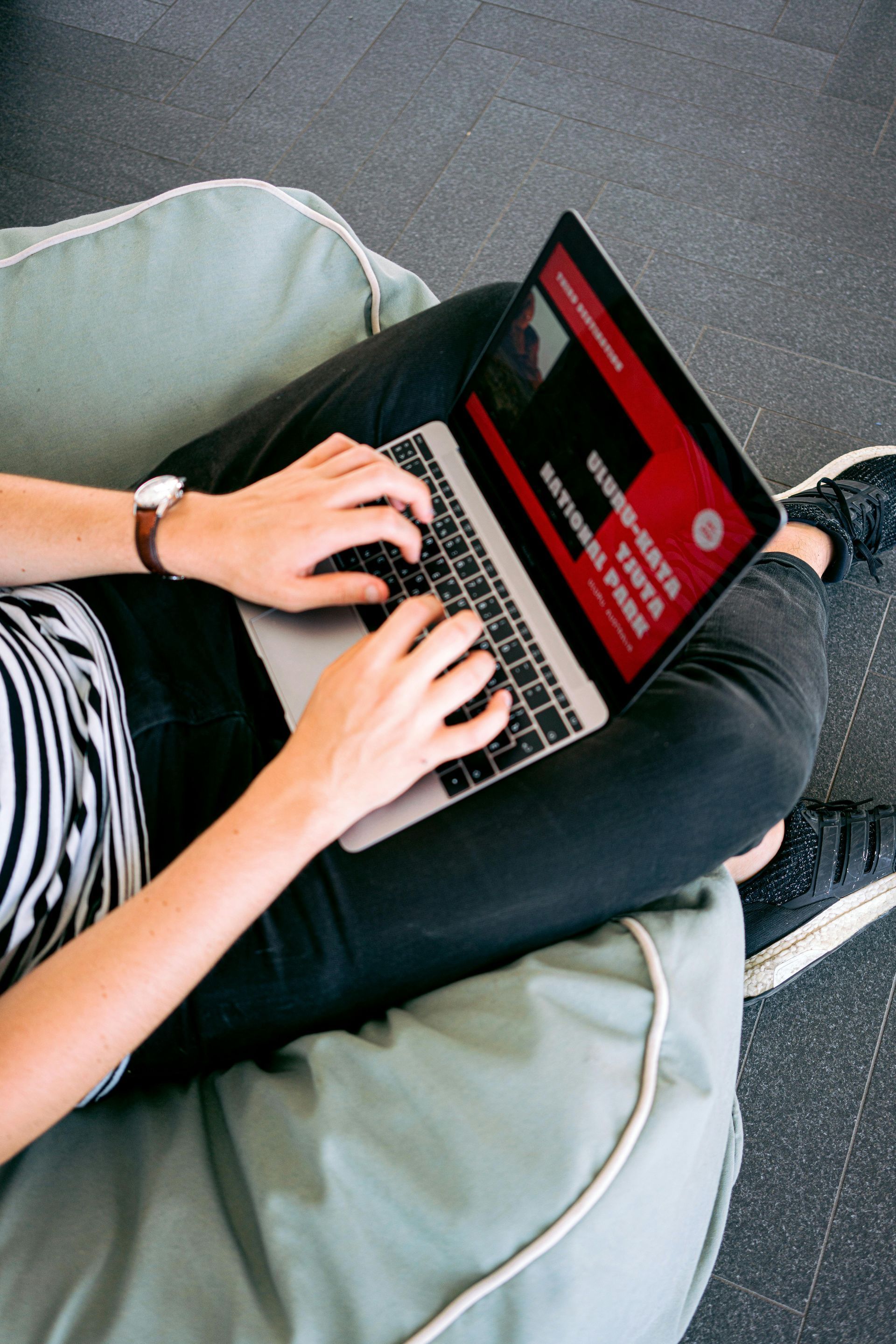 Person typing on a laptop with a red screen, sitting on a beanbag chair. They wear a watch, black pants, and black and white striped shirt.