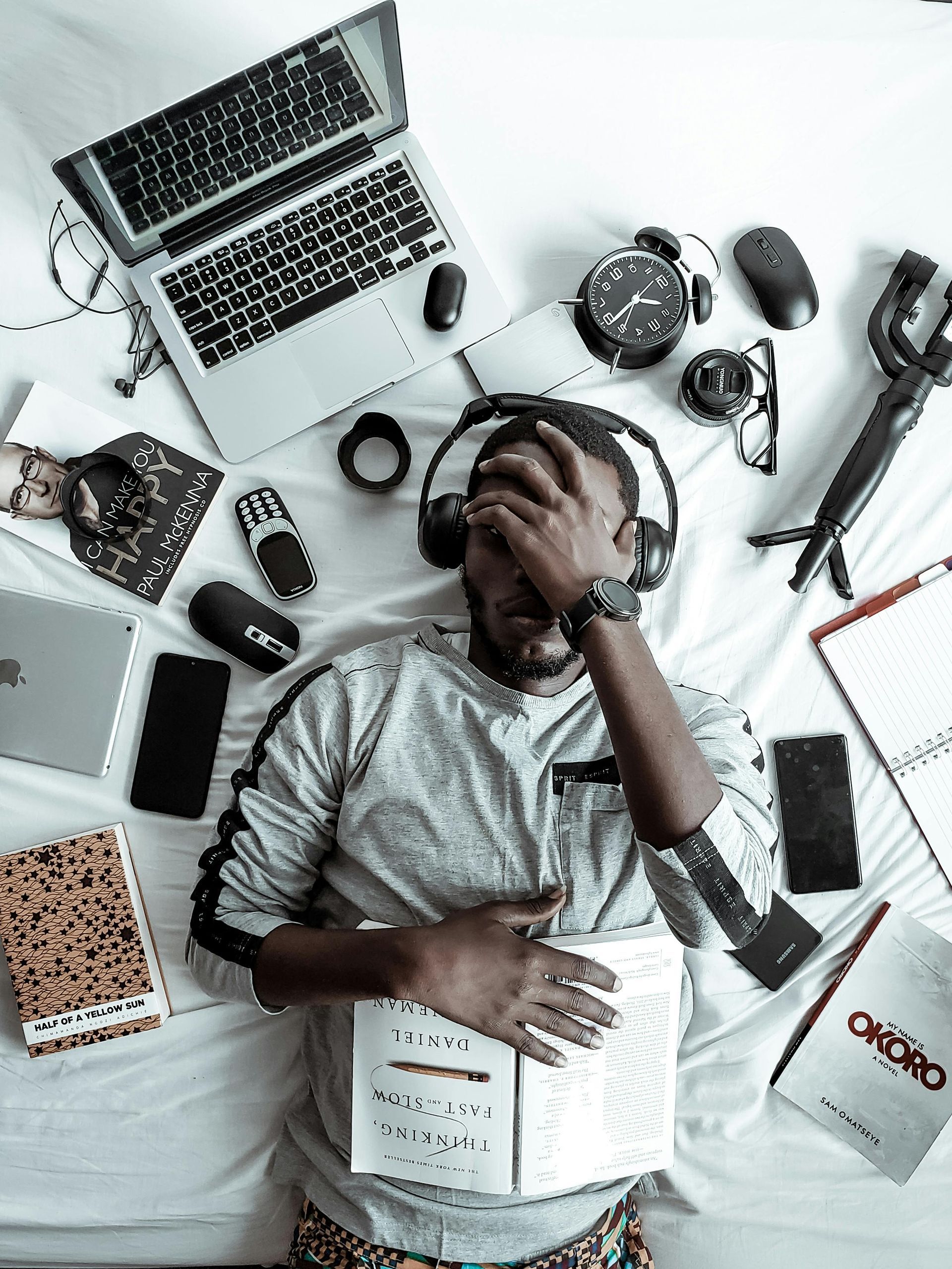Person lying on bed, surrounded by tech and books, looks stressed.