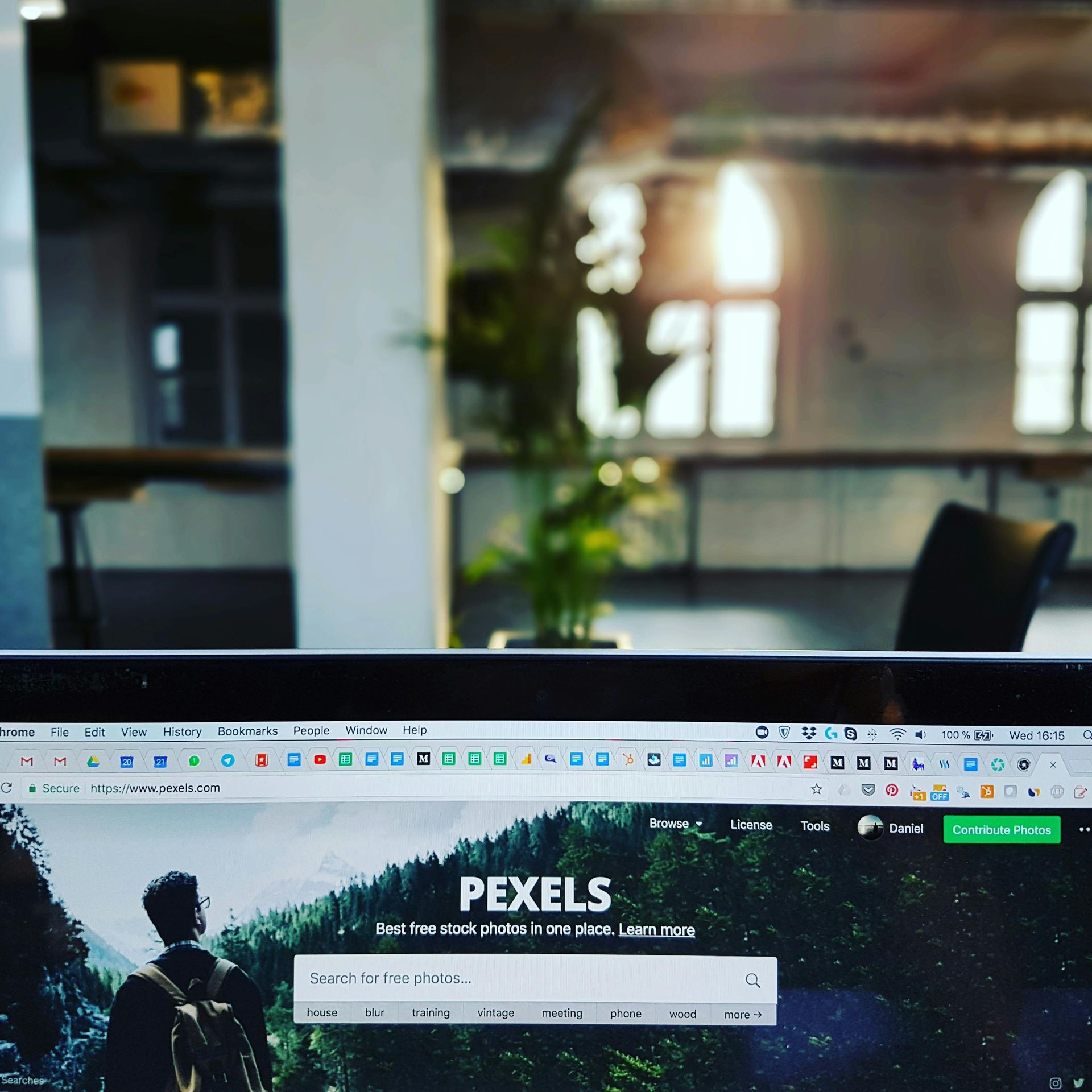 Laptop screen displaying the Pexels website, with a blurred office interior and sunlight in the background.
