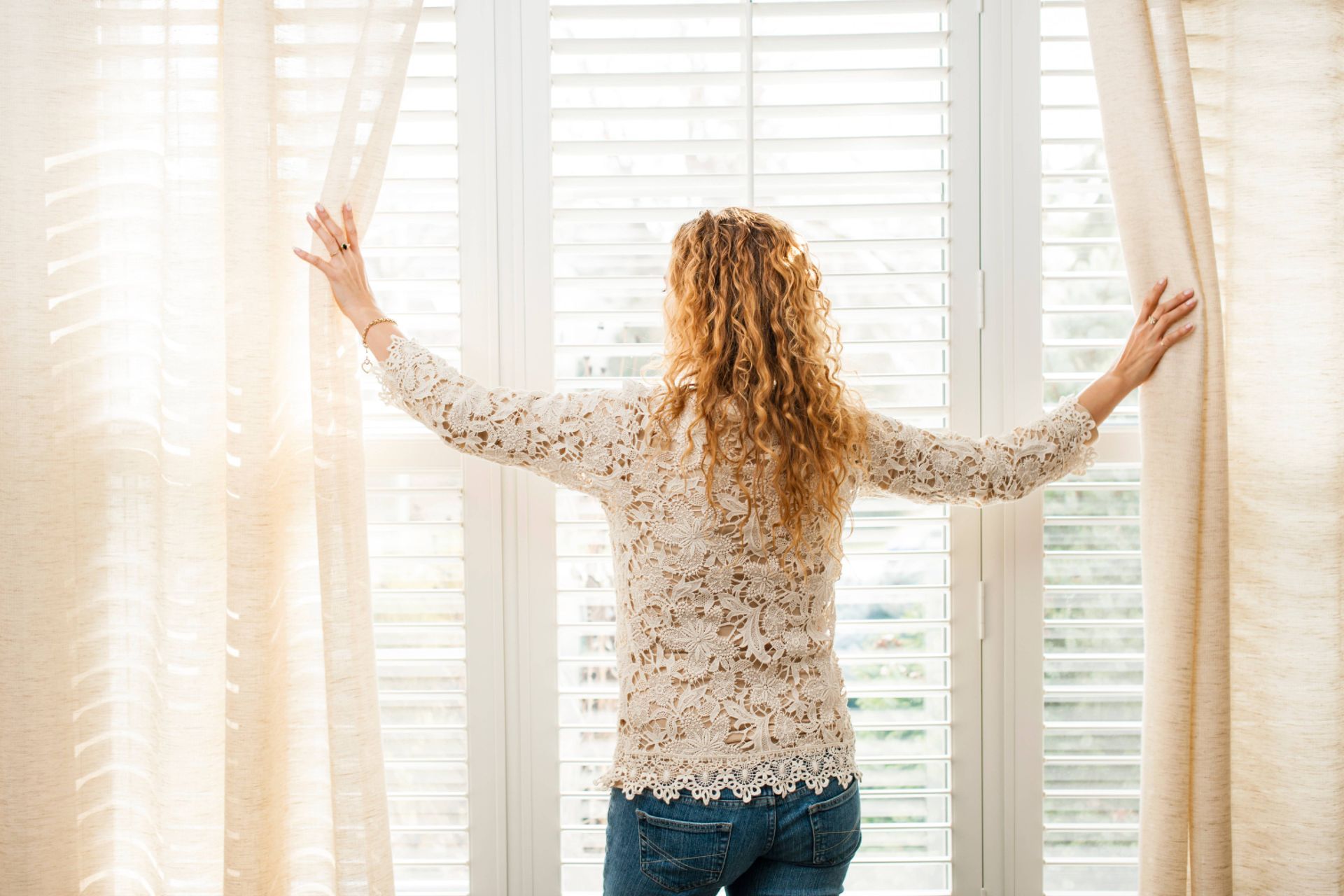 A woman opening her custom drapes to peer out of her window's custom shutters near Boynton Beach, FL