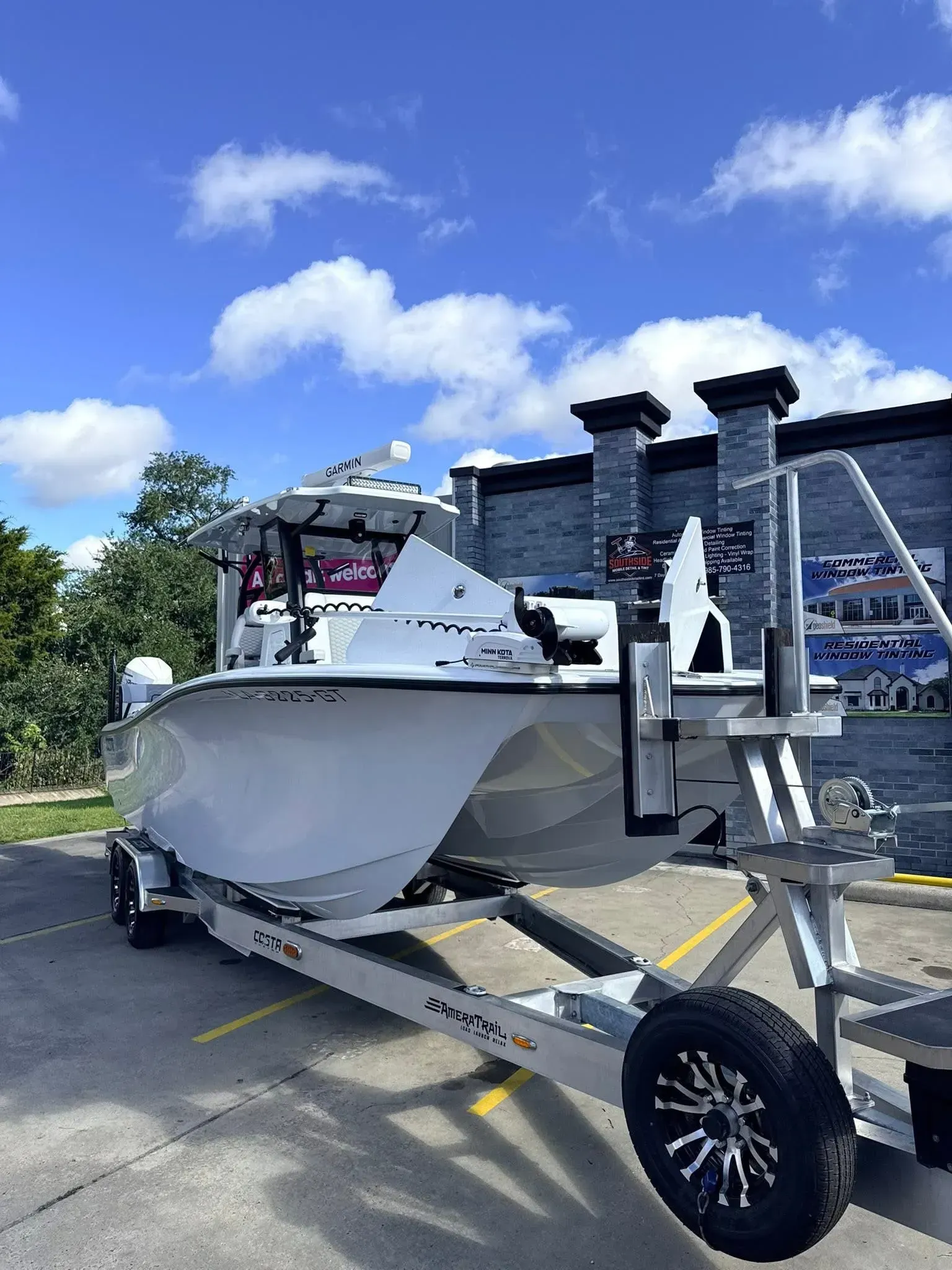 White boat on a trailer, parked in front of a building on a sunny day.