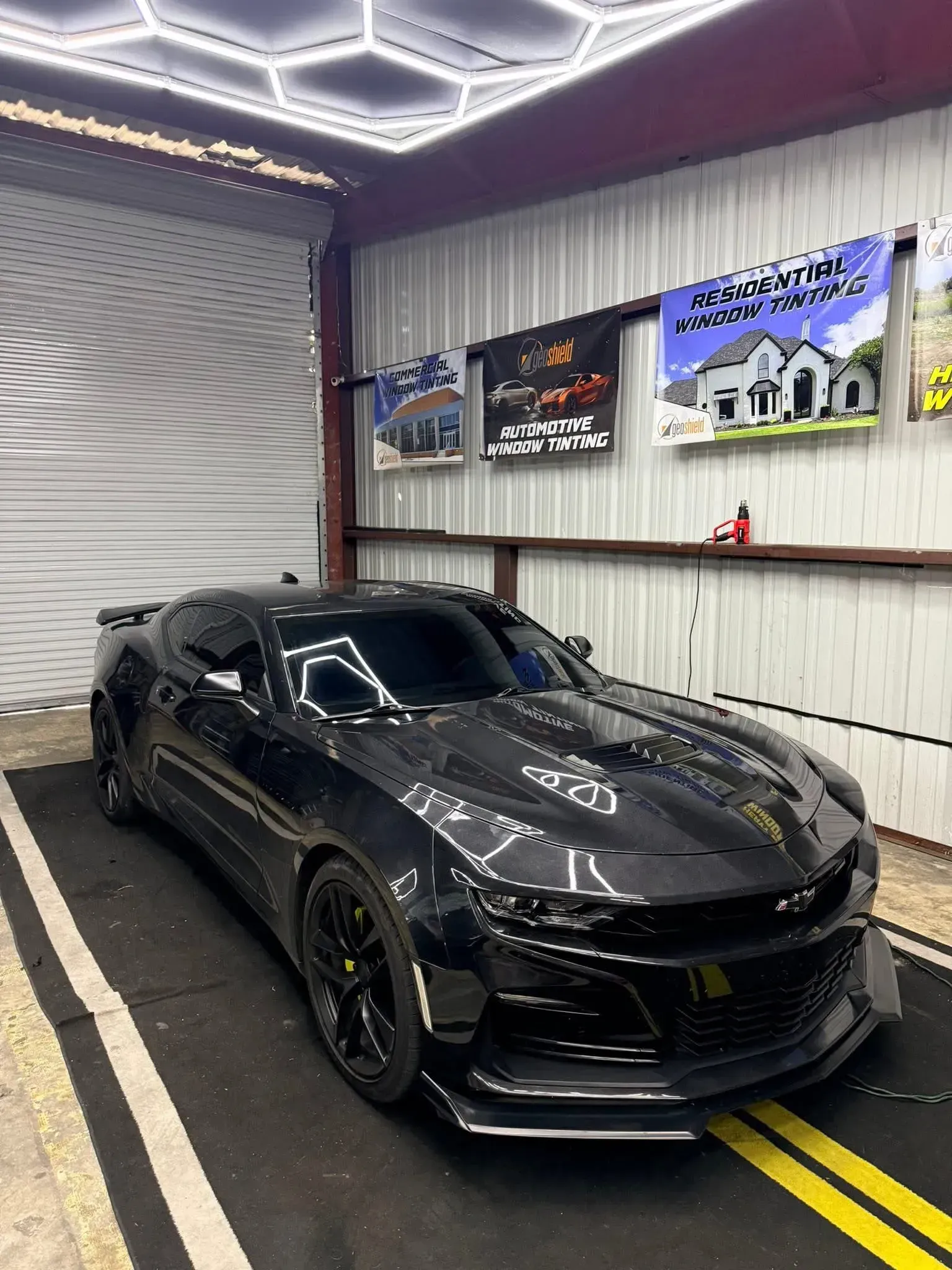 Black Chevrolet Camaro in a garage, black wheels, yellow line on the floor, promotional banners on the wall.