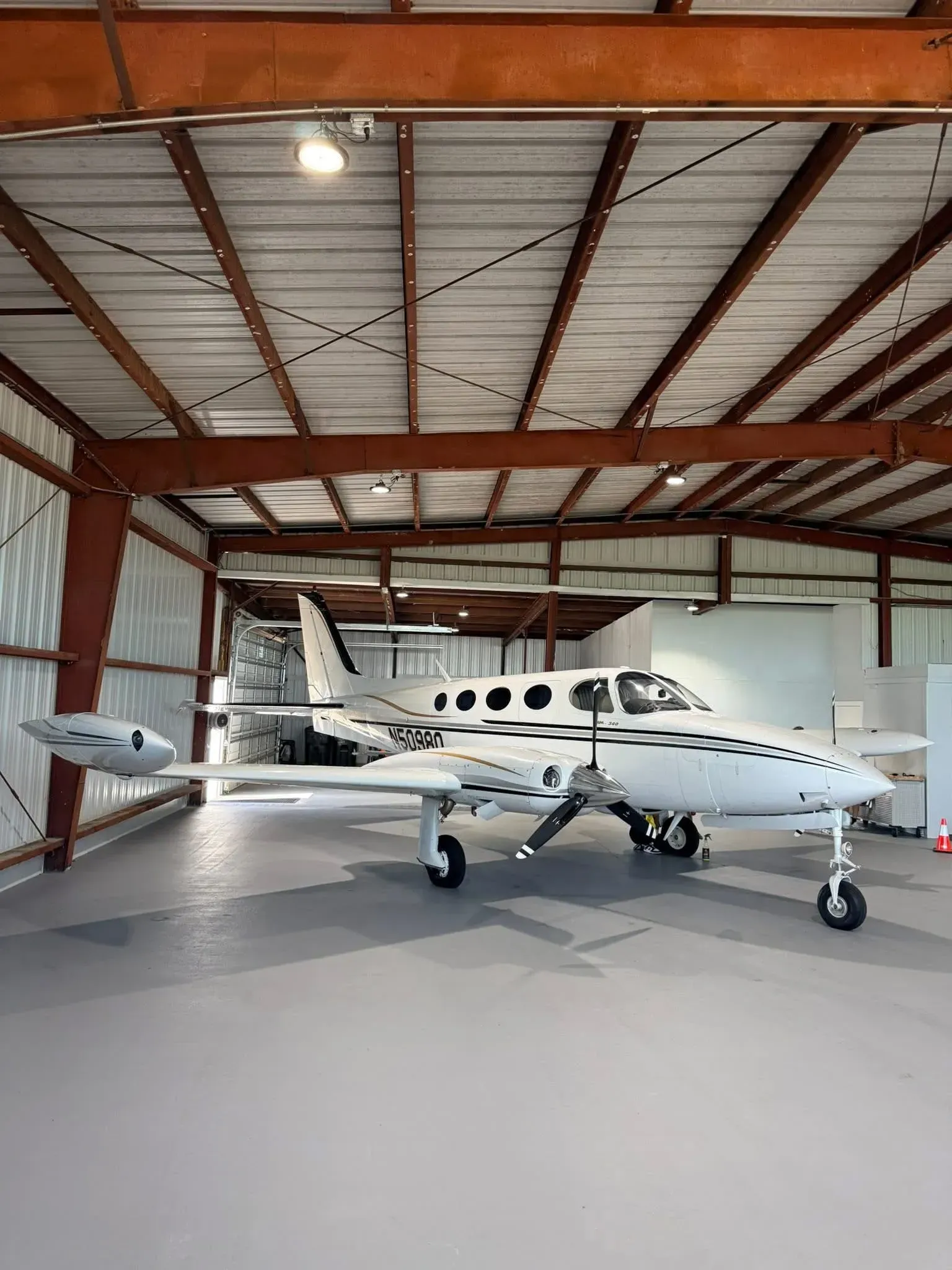 A small white airplane inside a hangar with brown metal beams and a gray floor.