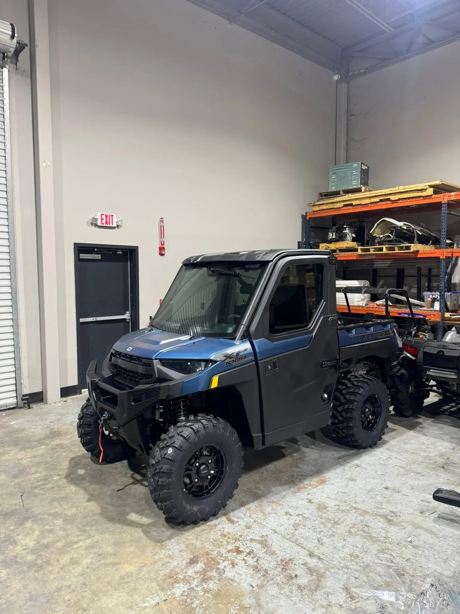 Blue utility task vehicle (UTV) parked inside a building. Black wheels, tires, and frame. Shelves and a closed door are in the background.