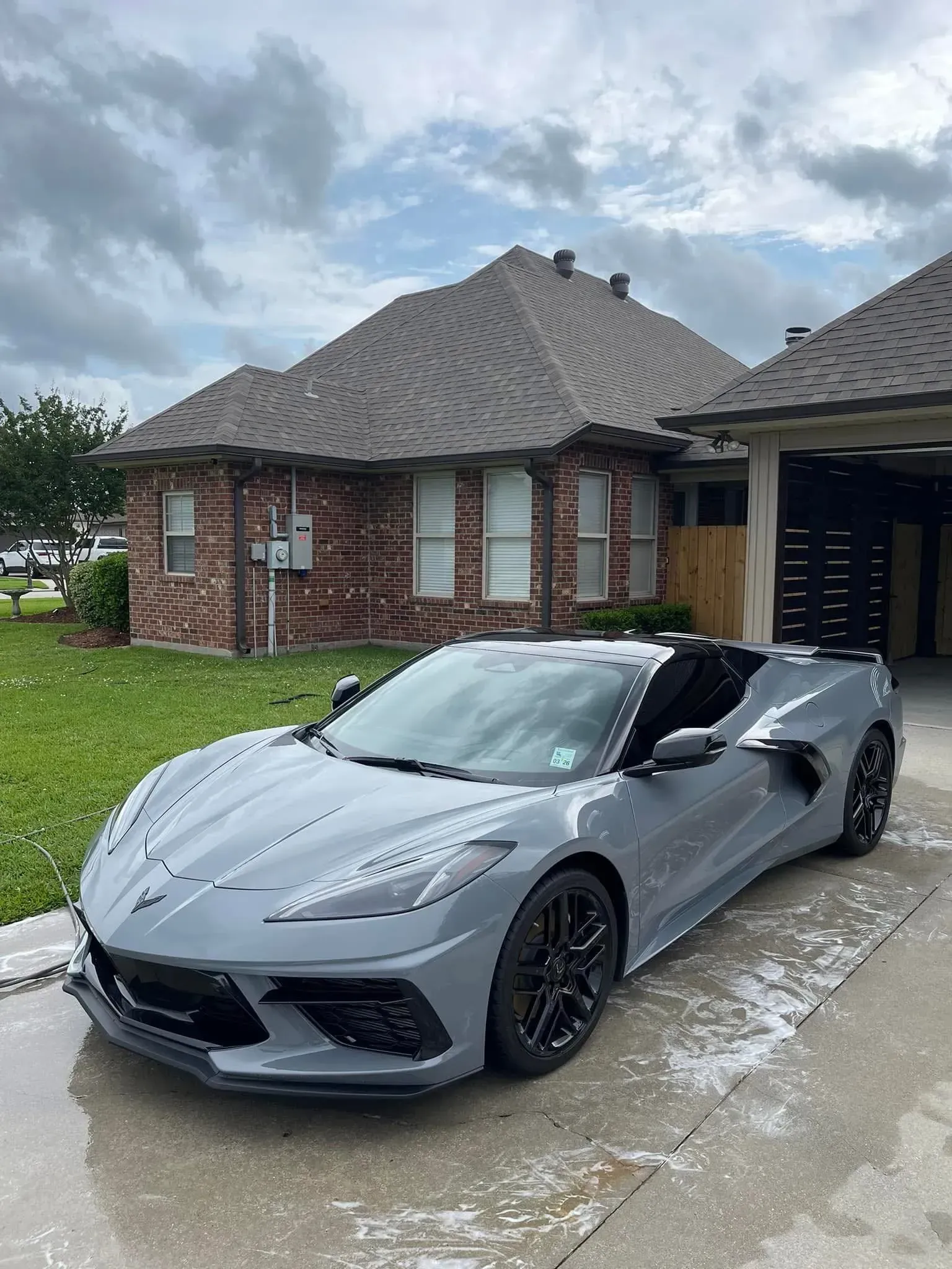Gray Chevrolet Corvette convertible parked in front of a brick house with a cloudy sky.