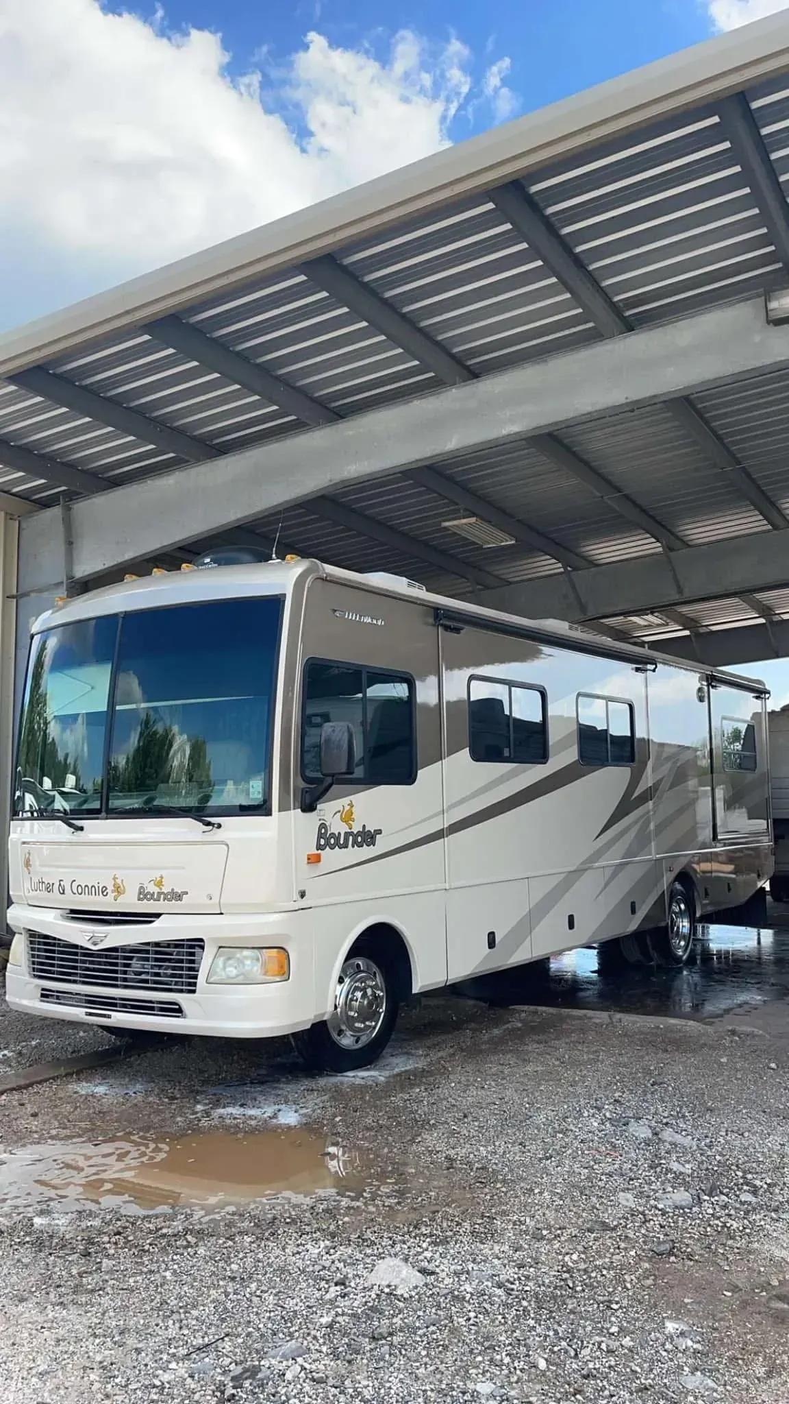 White and tan RV parked under a metal awning on a gravel surface.