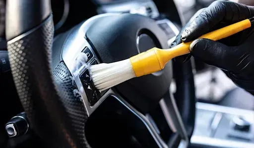Gloved hand using a yellow brush to clean buttons on a car's black steering wheel.