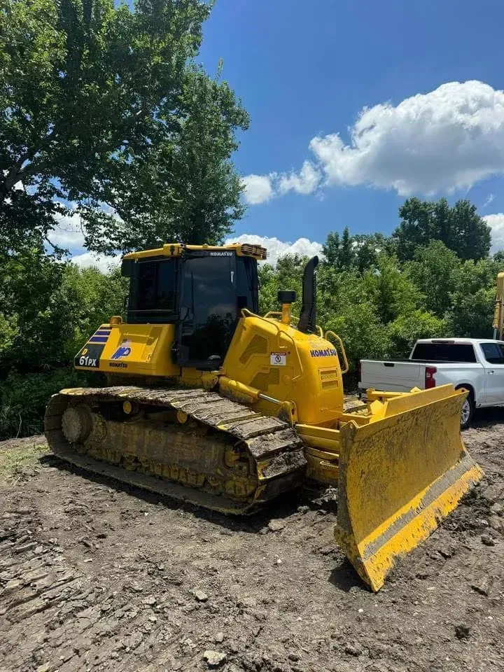 Yellow bulldozer with blade, parked on dirt, tree line, white truck, blue sky.