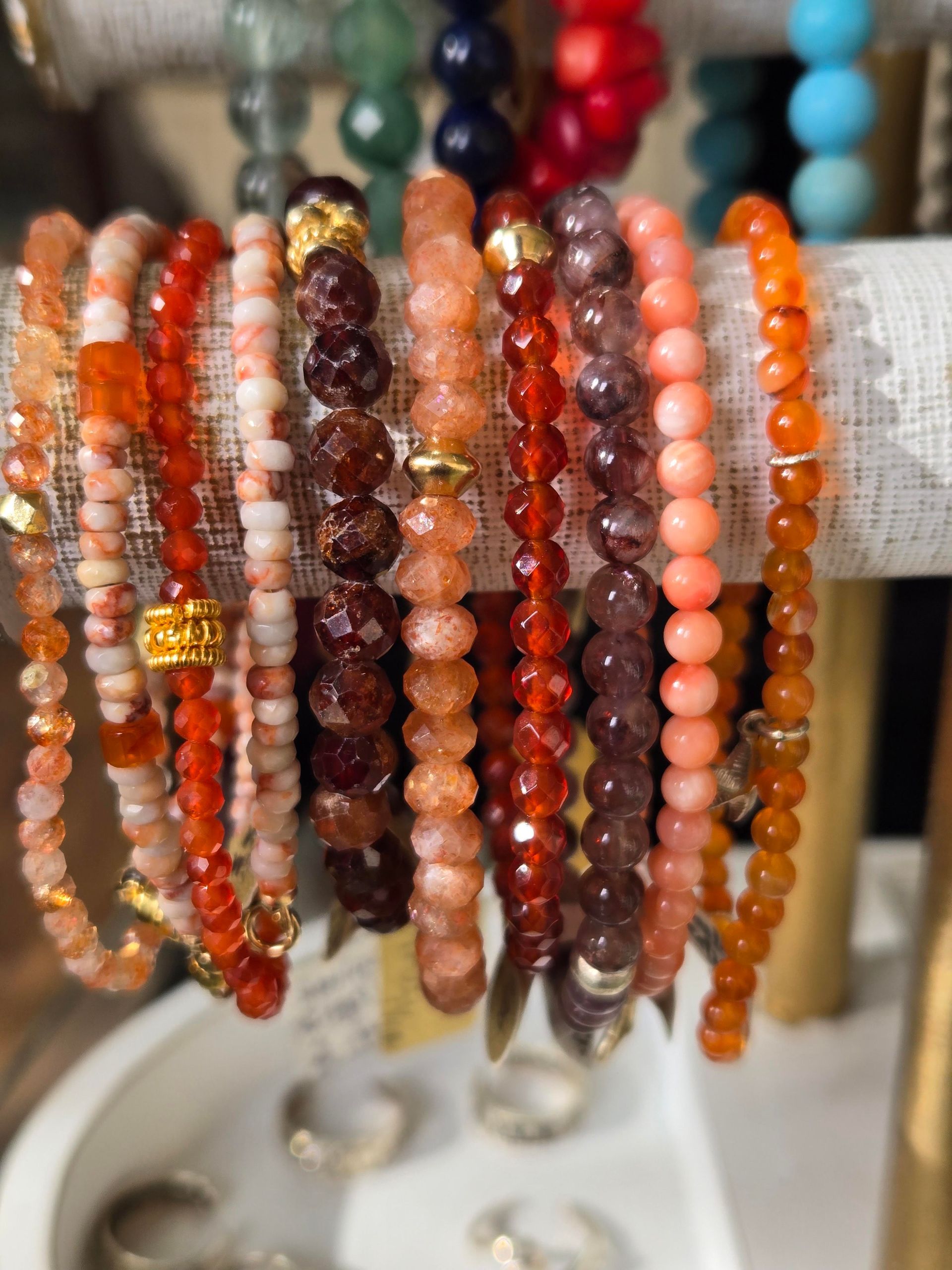 Jewelry designer sketching necklace designs on paper, with various gemstone necklaces and stones on a wooden table.