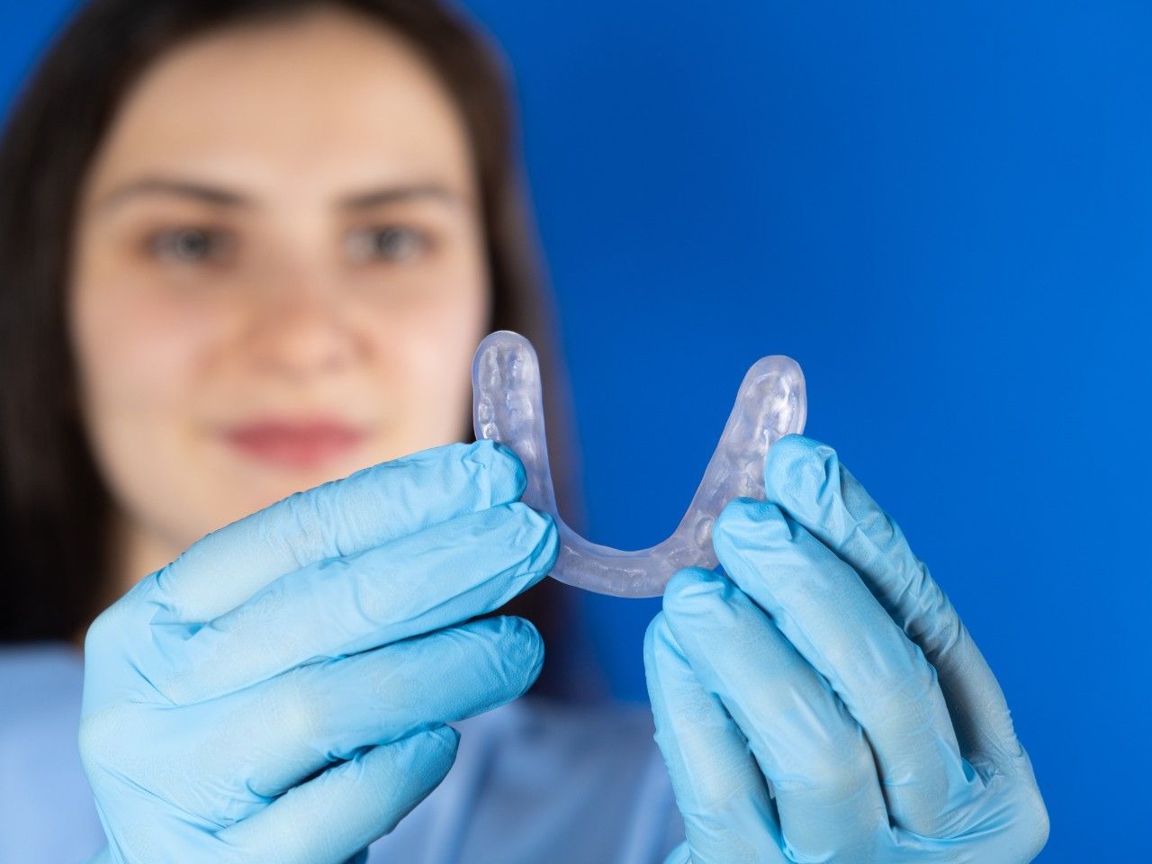 Persona con guantes azules sosteniendo un protector bucal transparente sobre un fondo azul.