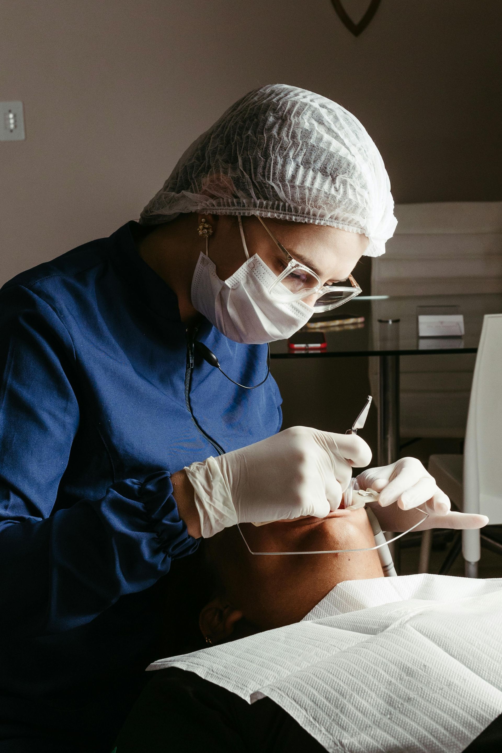 Dentista con uniforme azul, mascarilla y gorro, examinando la boca de un paciente con herramientas.