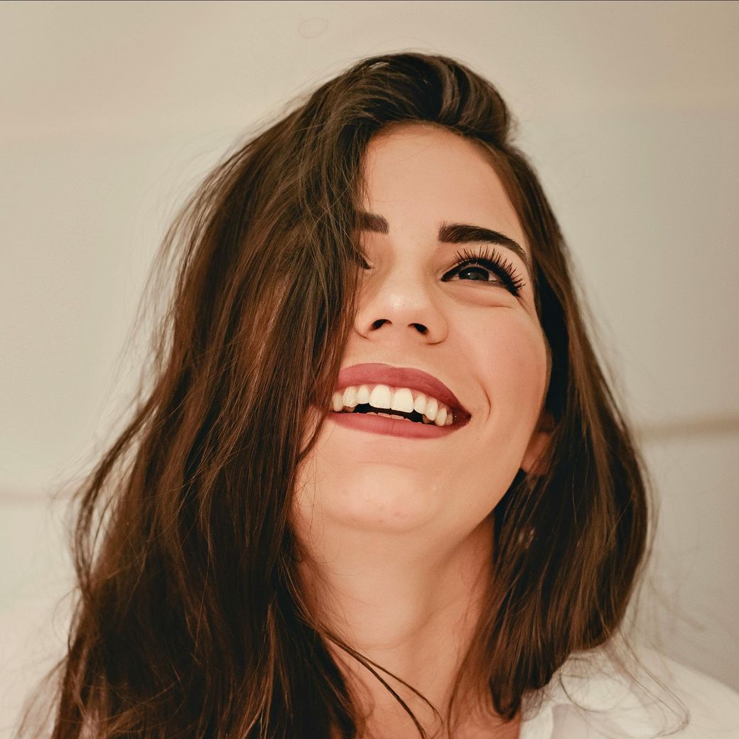Una mujer de cabello castaño sonríe ampliamente, mirando hacia arriba, vistiendo una camisa blanca.