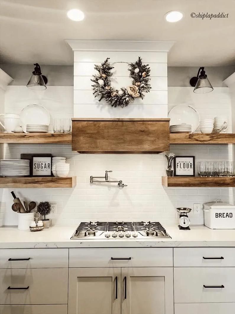 A kitchen with white cabinets , wooden shelves , a stove and a wreath on the ceiling.