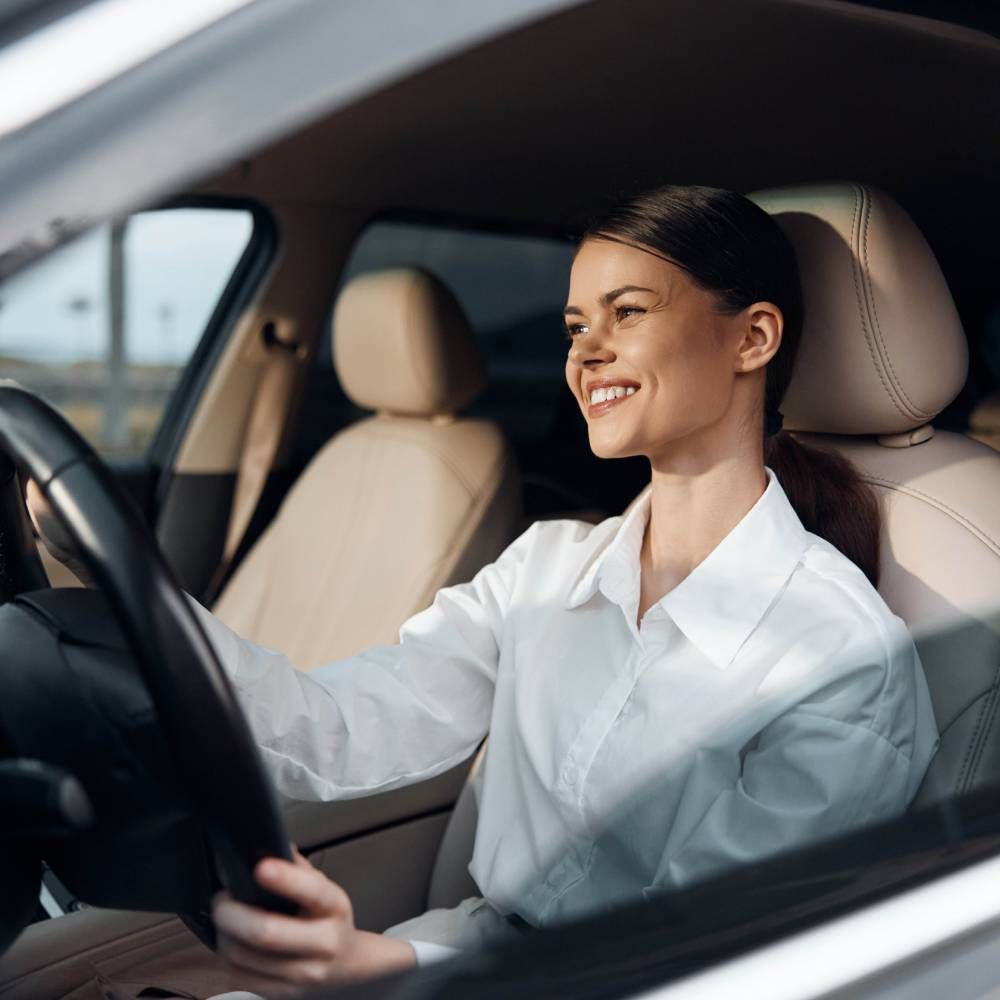 A woman in a white shirt is driving a car and smiling.