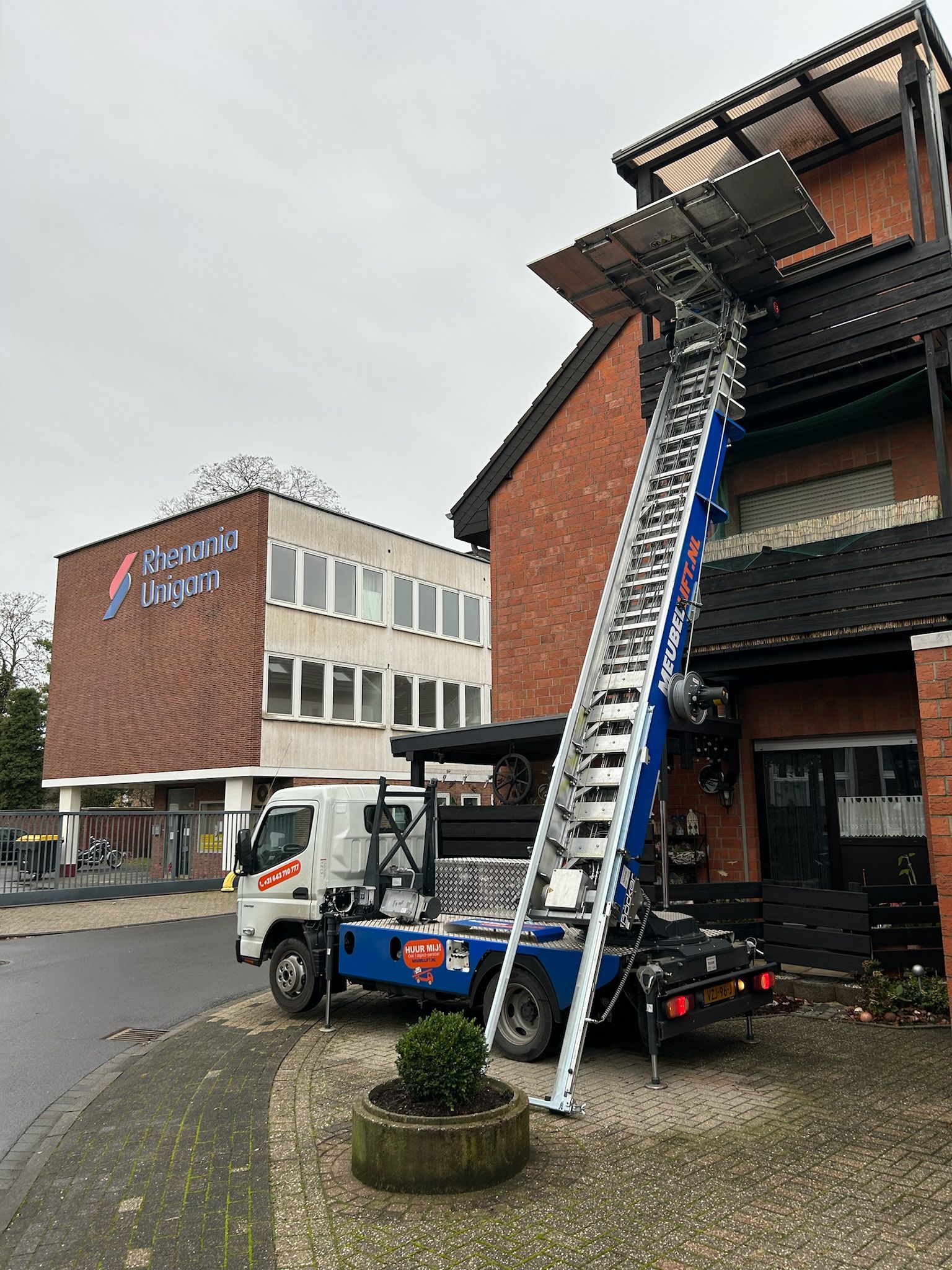 Een blauwe vrachtwagen met daaraan een ladder staat geparkeerd voor een bakstenen gebouw.
