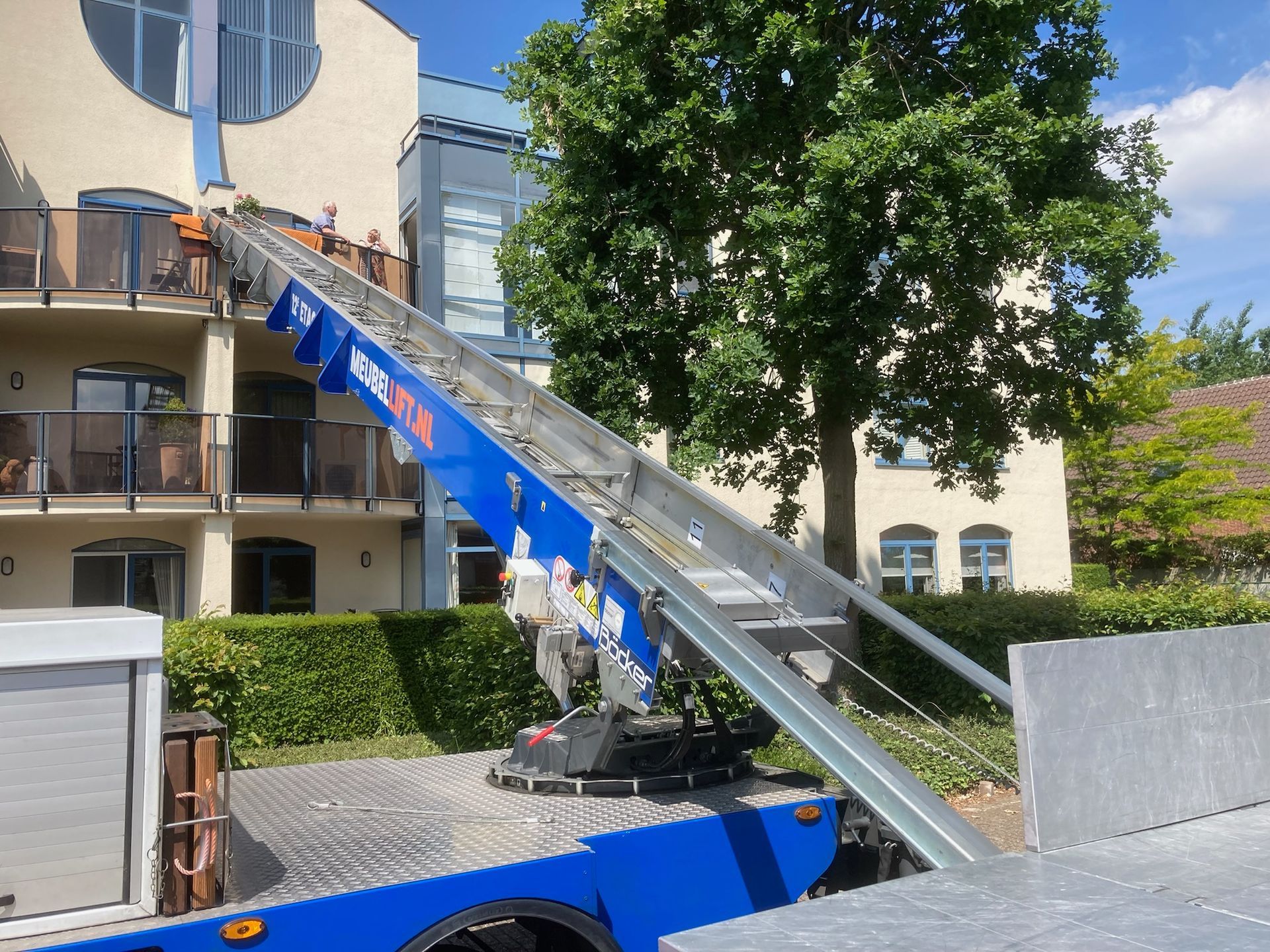 Een blauwe vrachtwagen met een ladder aan de achterkant staat geparkeerd voor een gebouw