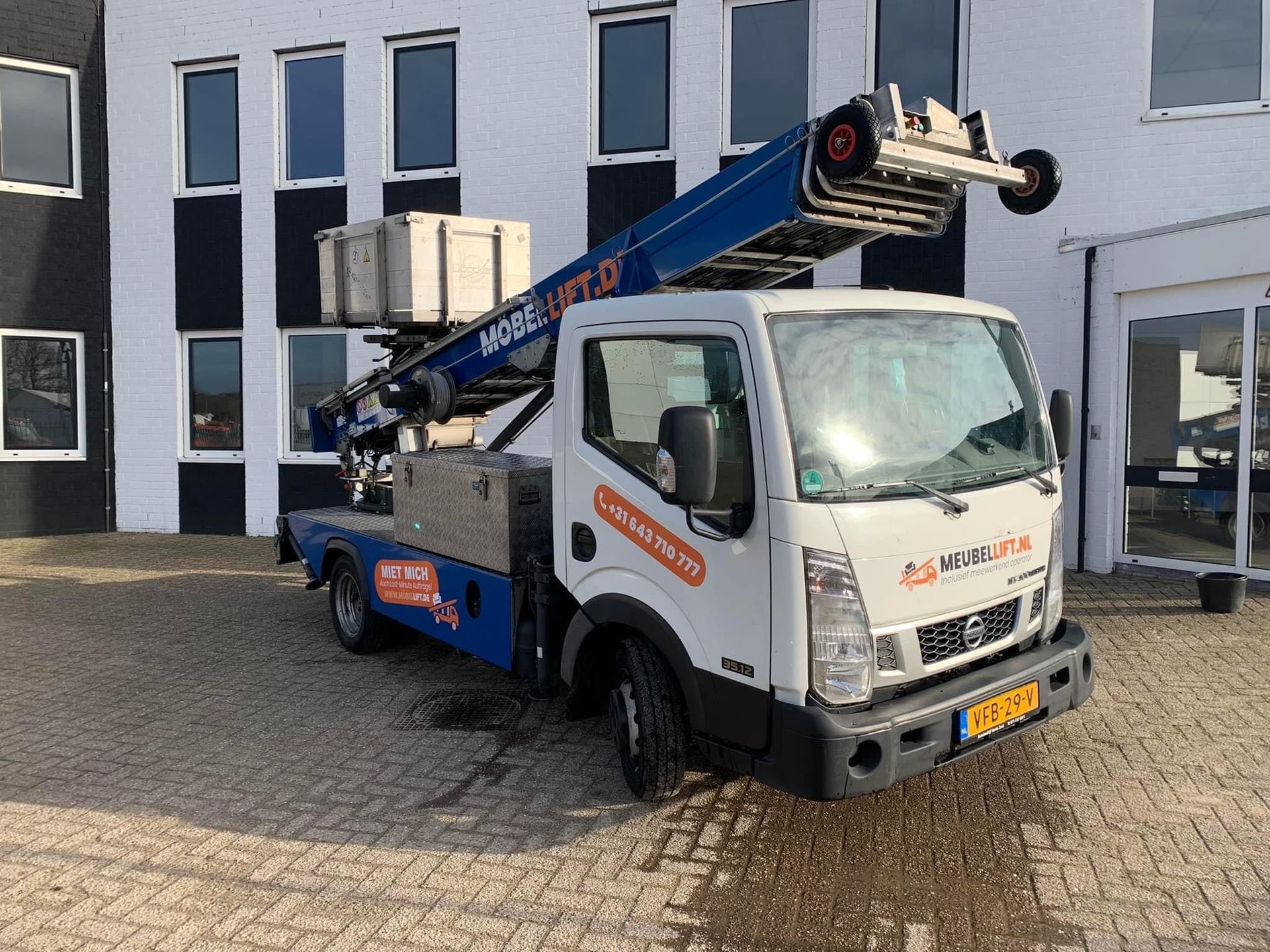 Een blauw-witte vrachtwagen met een ladder erop staat geparkeerd voor een gebouw.
