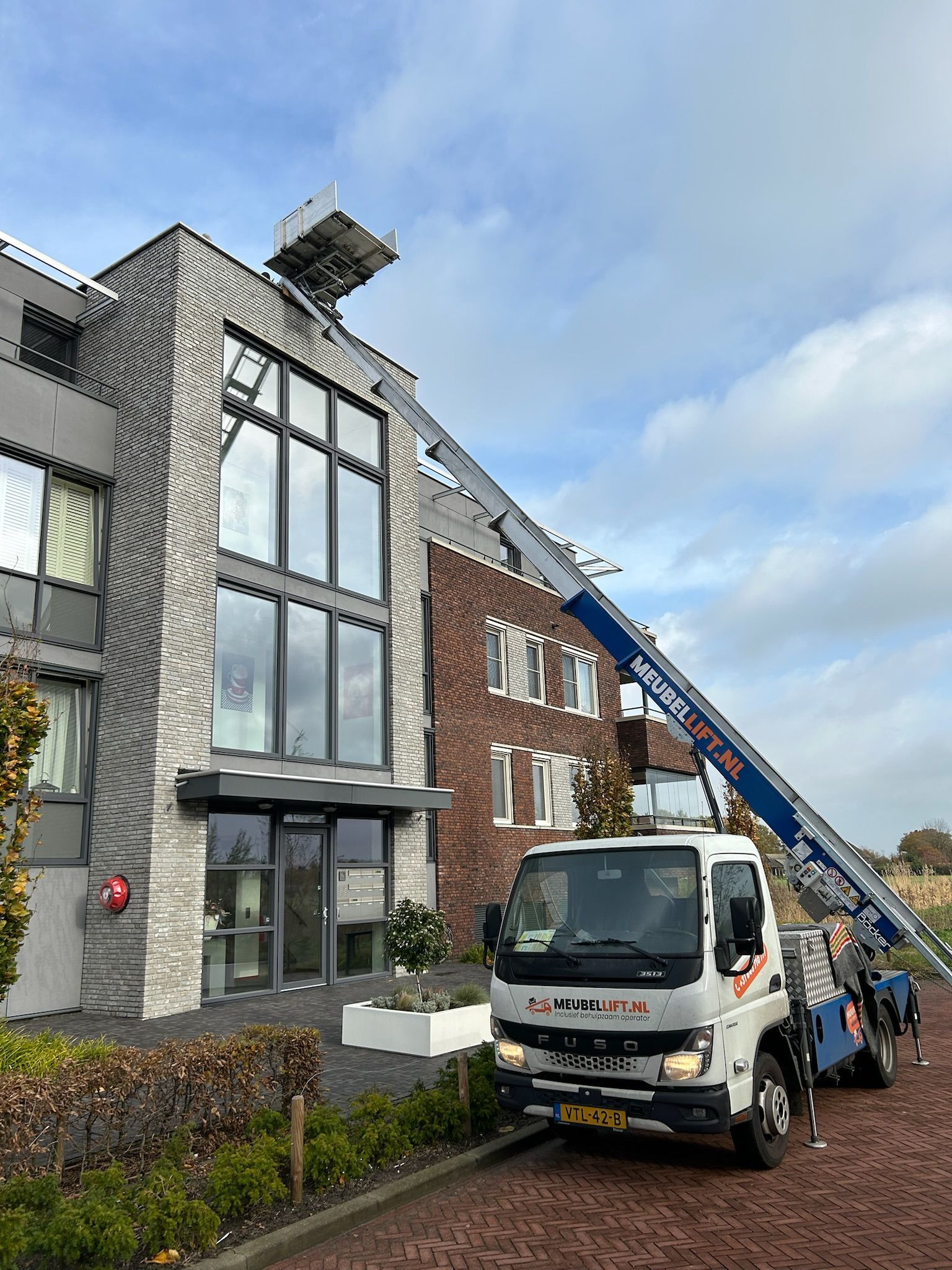 Een witte vrachtwagen met daarop een kraan staat geparkeerd voor een groot gebouw.