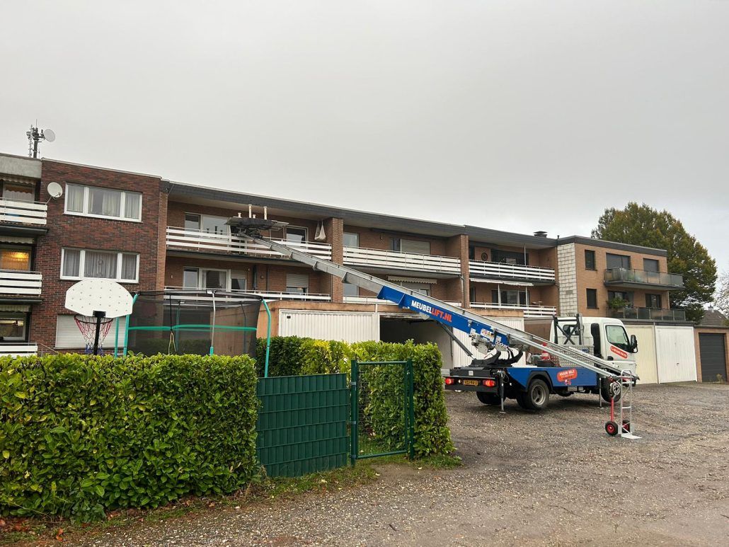 Een blauwe vrachtwagen met daaraan een ladder staat geparkeerd voor een gebouw.