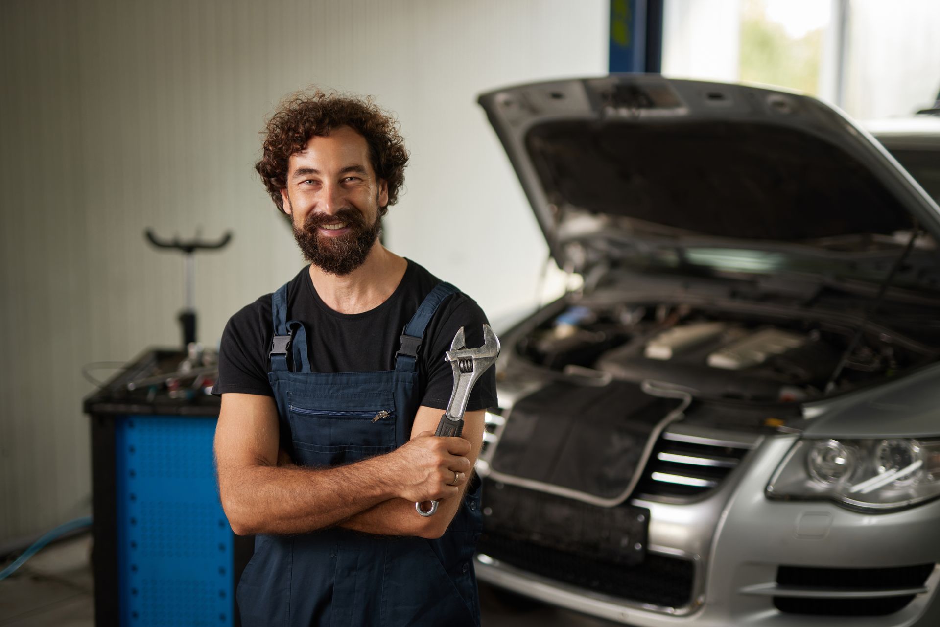 A car mechanic with a wrench in hand stands in front of a car with an open hood in an auto shop.